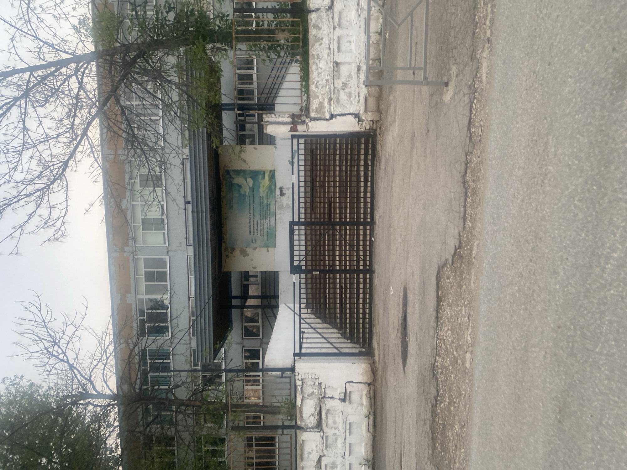 The image shows the exterior of a school building, identified by text on a mural as "Школа № 32 с углубленным изучением отдельных предметов" (School No. 32 with in-depth study of individual subjects) and located in Samara, Russia. The building is a multi-story structure with a gray facade and numerous windows. In front of the building, there are metal gates and a set of stairs leading up to the entrance. The area in front of the school is paved with asphalt and shows signs of wear and tear, with potholes and cracks visible. Bare trees with intricate branches frame the building. A mural is affixed to the wall to the right of the entrance, featuring a sky scene with clouds and birds. The overall impression is of a utilitarian and somewhat aged educational institution.