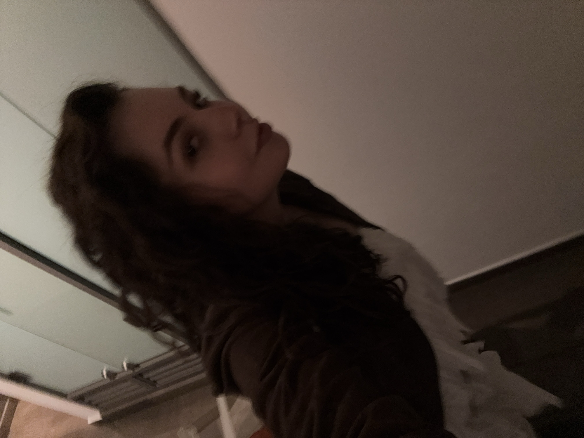A medium shot depicts a woman with long, dark, wavy hair and light skin. She's wearing a dark brown garment over a white top. The background consists of a light-colored wall and what appears to be a window frame. The photograph seems to be taken at an angle, looking slightly upward at the subject. The setting cannot be verfied to be Mexico City, Mexico.