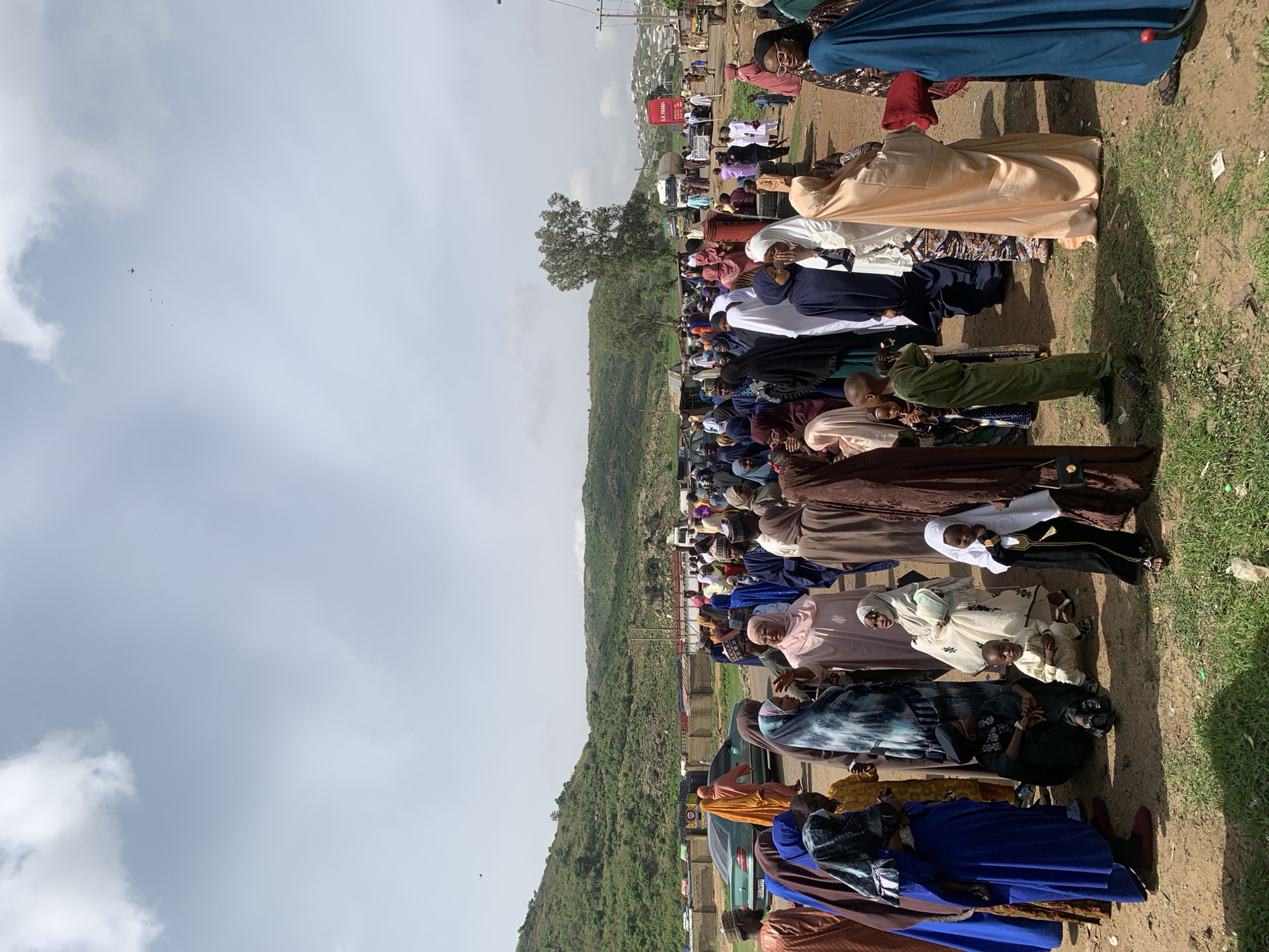 A large group of people are gathered in an open area in Naraguta Hausawa, Nigeria. Many are sitting on mats, seemingly praying or engaged in religious observance. Individuals are wearing a variety of colorful, traditional clothing. The terrain is mostly dirt with patches of grass. A hill is visible in the background. Some vehicles are parked nearby. A utility pole can be seen on the edge of the crowd. The sky is overcast with scattered clouds.