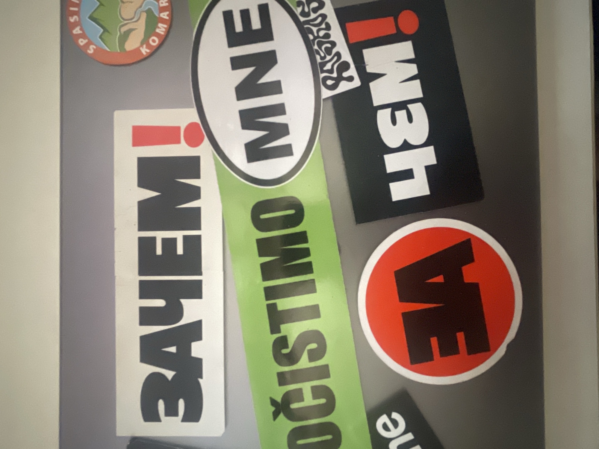 The image displays a collection of seven stickers affixed to a flat, gray surface, possibly a wall or cabinet. The stickers are oriented vertically, suggesting the image itself is rotated 90 degrees counter-clockwise.
The visible stickers include:
1. A circular sticker in the top left corner, featuring a stylized landscape of mountains and a river, with partially visible text around the edge that reads "SPASIJA KOMARA".
2. A rectangular white sticker below it, bearing large black capital letters that spell "ЗАЧЕМ!" (ZACHEM!, meaning "Why!"), accompanied by a red exclamation mark.
3. An oval white sticker in the top center, with black capital letters reading "MNE", likely an abbreviation for Montenegro.
4. A long rectangular light green sticker extending diagonally across the center, with black capital letters spelling "OČISTIMO" (meaning "Let's clean" in several South Slavic languages).
5. A rectangular black sticker in the top right, with white, stylized, vertically oriented text that appears to be "!WЭН" (possibly "!MEN" or "!WEN", with 'Э' being a Cyrillic letter for E). It also has a red exclamation mark.
6. A circular red sticker in the bottom right, featuring two large black capital letters "AE".
7. A small, partially visible sticker at the very bottom, showing black letters "ne".
The scene is static, featuring only these decorative or informational stickers. There is no activity taking place, and no specific time of day or weather can be discerned. The stickers use a mix of Latin and Cyrillic alphabets.