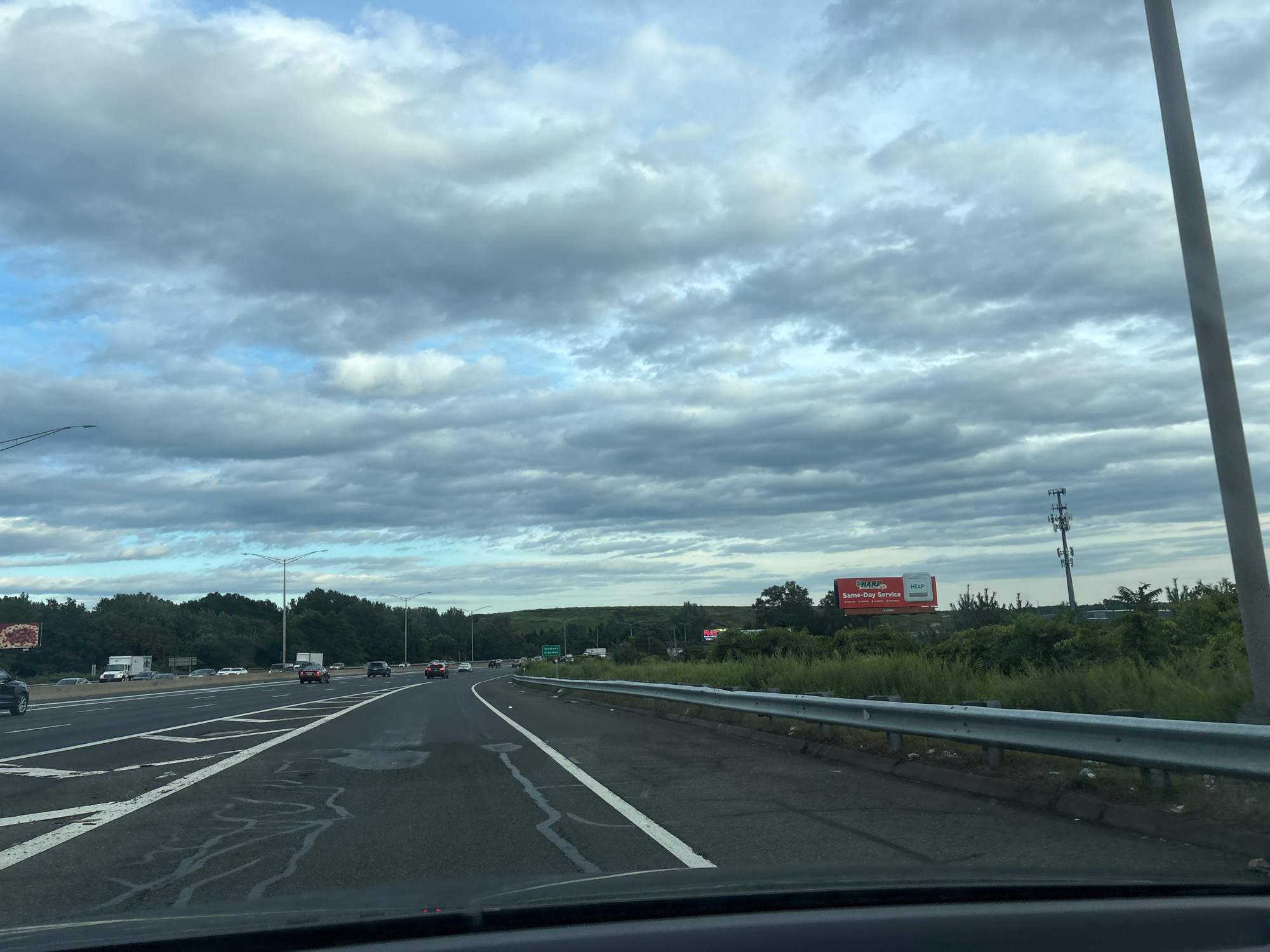 The image captures a view from a vehicle driving on a multi-lane highway, likely in or around Hartford, Connecticut, USA. The sky is overcast with dramatic, layered clouds. The highway itself appears wet in patches, possibly from recent rain. Lanes are marked with white lines, and a metal guardrail separates the road from a grassy embankment on the right. Trees and foliage line the sides of the highway. Several vehicles are visible on the road, including cars and a truck. In the distance, billboards and a cell tower can be seen. One billboard prominently displays "HARP Same-Day Service HELP". The overall impression is one of travel on a cloudy day.