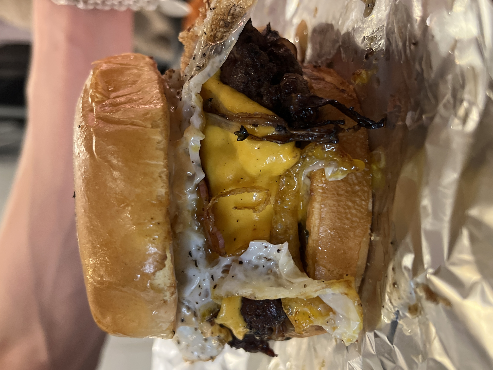 A close-up shot of a burger in a foil wrapper. The burger consists of a glazed bun, a beef patty, melted yellow cheese, fried onions, and a fried egg. The foil wrapper is crumpled around the burger. The location is unknown.
