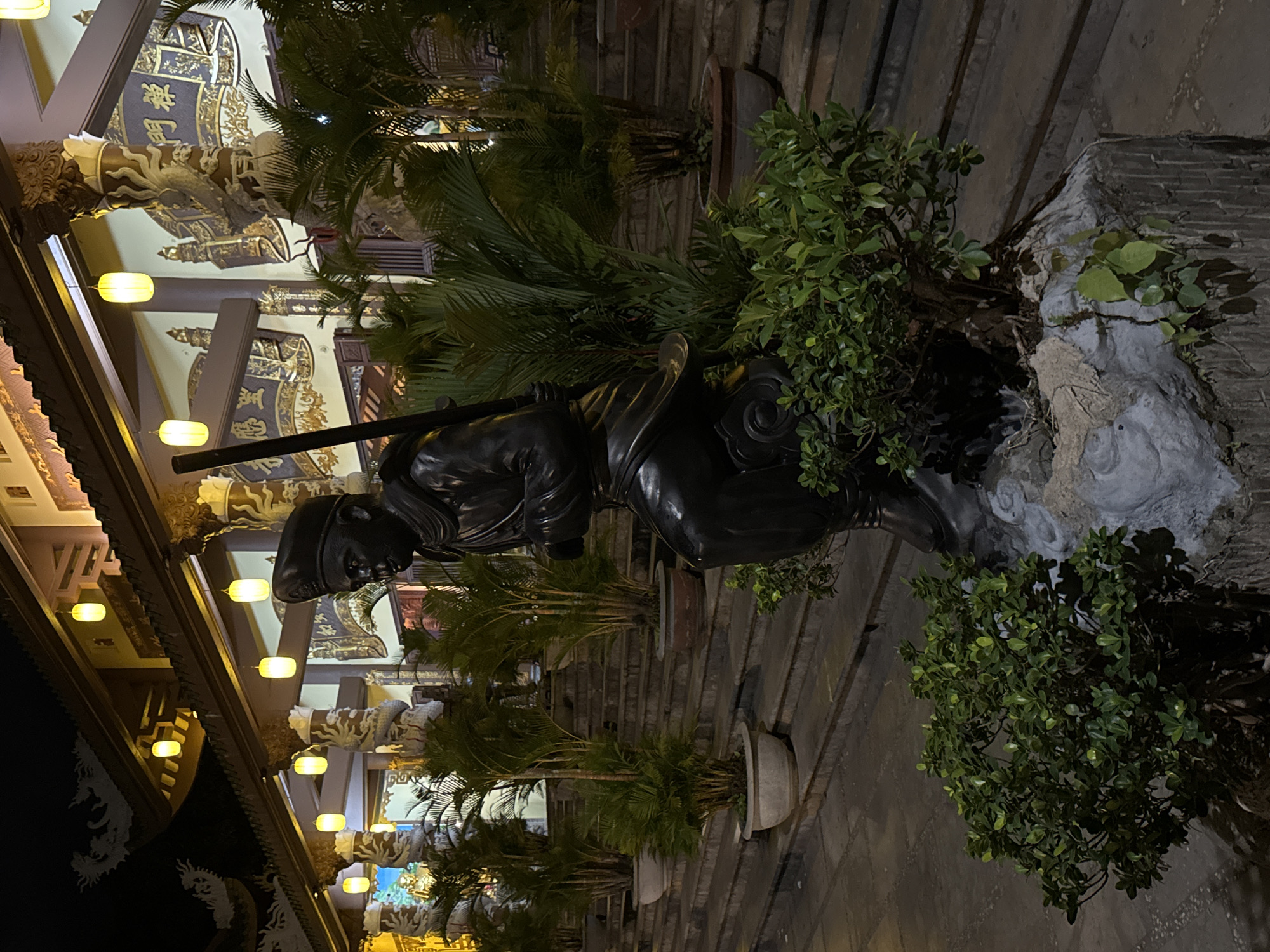An outdoor nighttime photograph captures a dark bronze statue of a seated figure, possibly a monk or a warrior, holding a staff, positioned in front of a temple or religious building with traditional Vietnamese architectural elements. The statue is situated on a raised stone platform adorned with potted plants, some of which are palm trees. The background features a multi-tiered staircase leading up to the building, which is illuminated by strings of yellow lanterns hanging from the eaves. The building's facade is decorated with intricate carvings, including dragons and ornate calligraphy. The overall scene suggests a serene and spiritual atmosphere, typical of a Buddhist temple. While specific details about the statue's identity are not explicitly provided, its attire and pose, combined with the temple setting, hint at its religious or cultural significance. The image appears to have been taken in Da Nang, Vietnam, a city known for its numerous temples and pagodas.