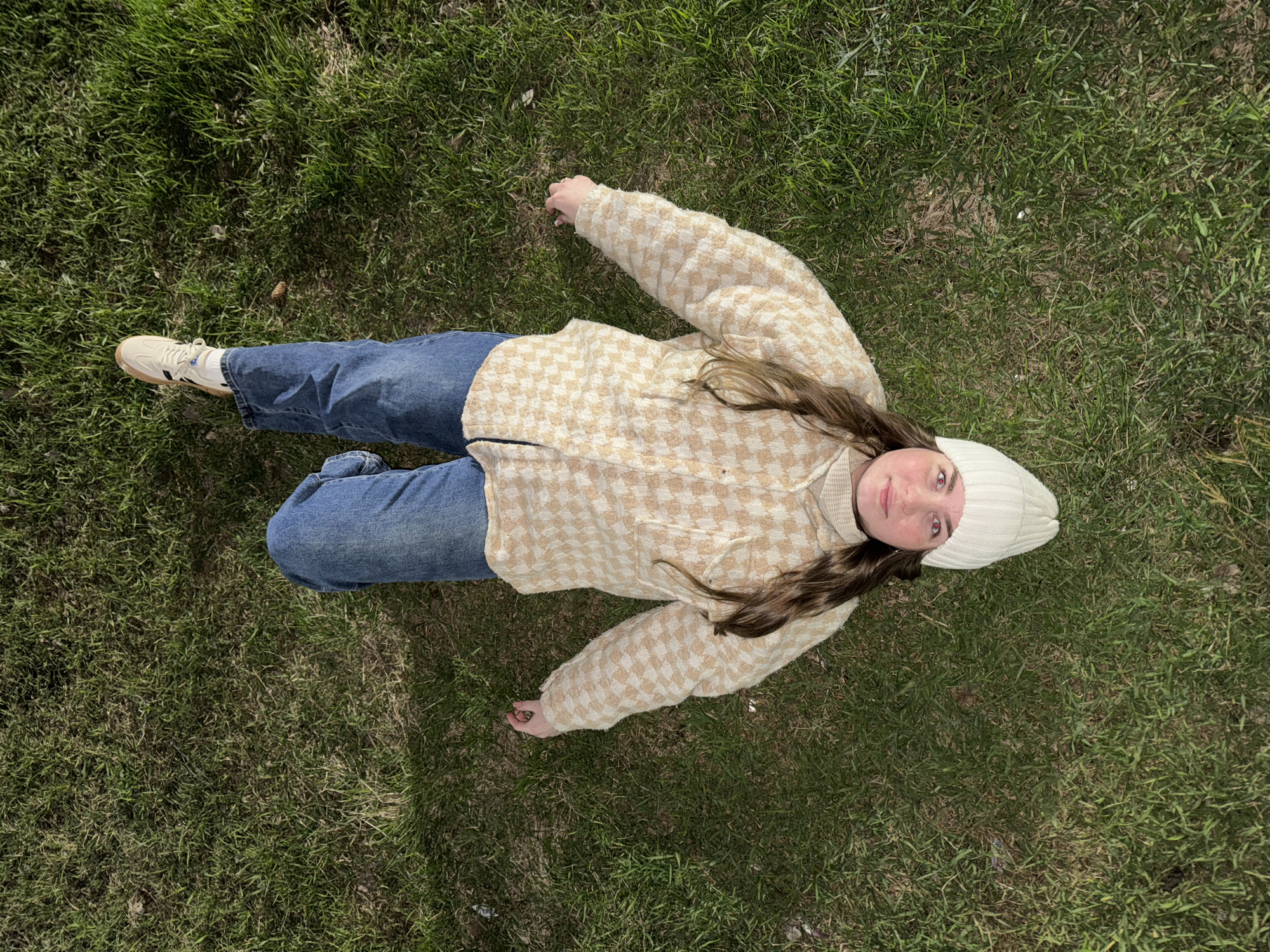 An aerial, full-body shot captures a young individual lying on their back on a vibrant green lawn in Chaikovsky, Russia. The person is dressed in a casual, warm outfit, including a light-colored knit beanie hat, a beige and cream checkered (houndstooth-like) jacket or cardigan, blue jeans, and light-colored sneakers with black stripes. Their long, brown hair spreads out on the grass around their head.
The individual is looking directly up at the camera with a slight, calm expression, their eyes showing a red-eye effect from the flash or lighting. One arm is extended slightly upwards, while the other is bent, resting near their side. Their legs are bent at the knees, with their feet visible at the bottom left of the frame.
The scene is an outdoor setting, likely a park or field, characterized by lush green grass with some sparse patches. The bright, overhead sunlight casts clear shadows, indicating a sunny daytime. There is no visible text in the image. The overall impression is one of relaxation or a posed moment amidst nature.