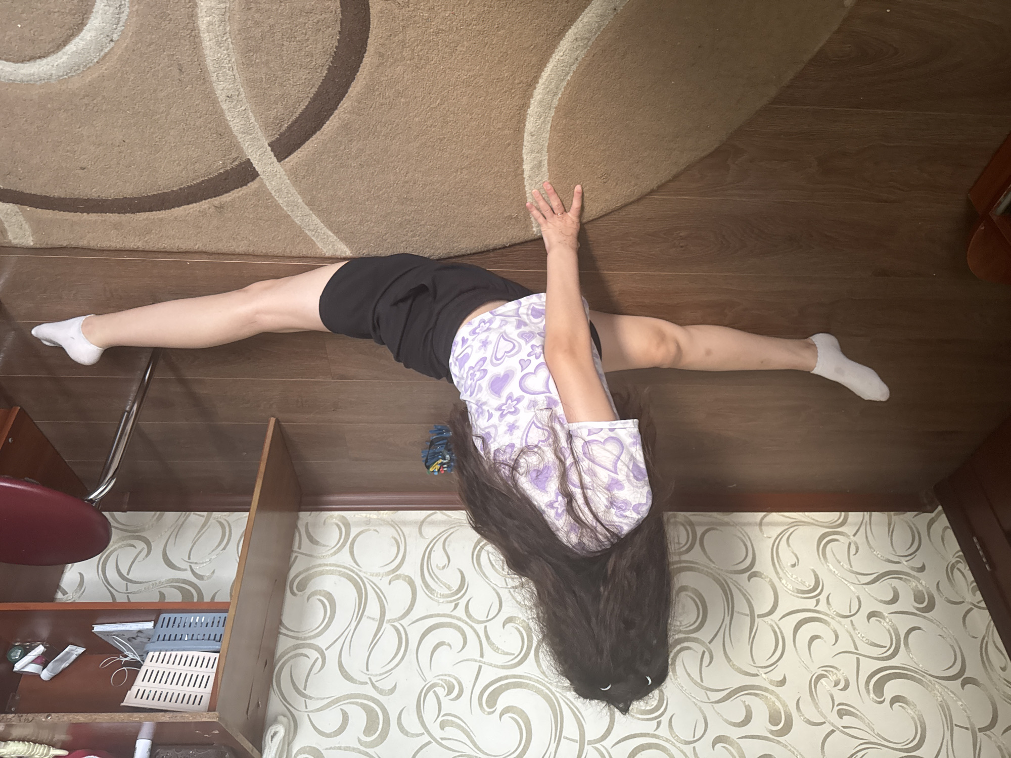 A woman with long, dark hair is in the middle of a split on a wooden floor. She is wearing a purple and white patterned t-shirt and black shorts. Her left leg is extended to the left and her right leg is extended to the right. Her right hand is resting on a tan rug with a circular pattern. In the background, there is a wall with a decorative wallpaper pattern and a wooden desk with various items on it, including a chair with a red seat.
