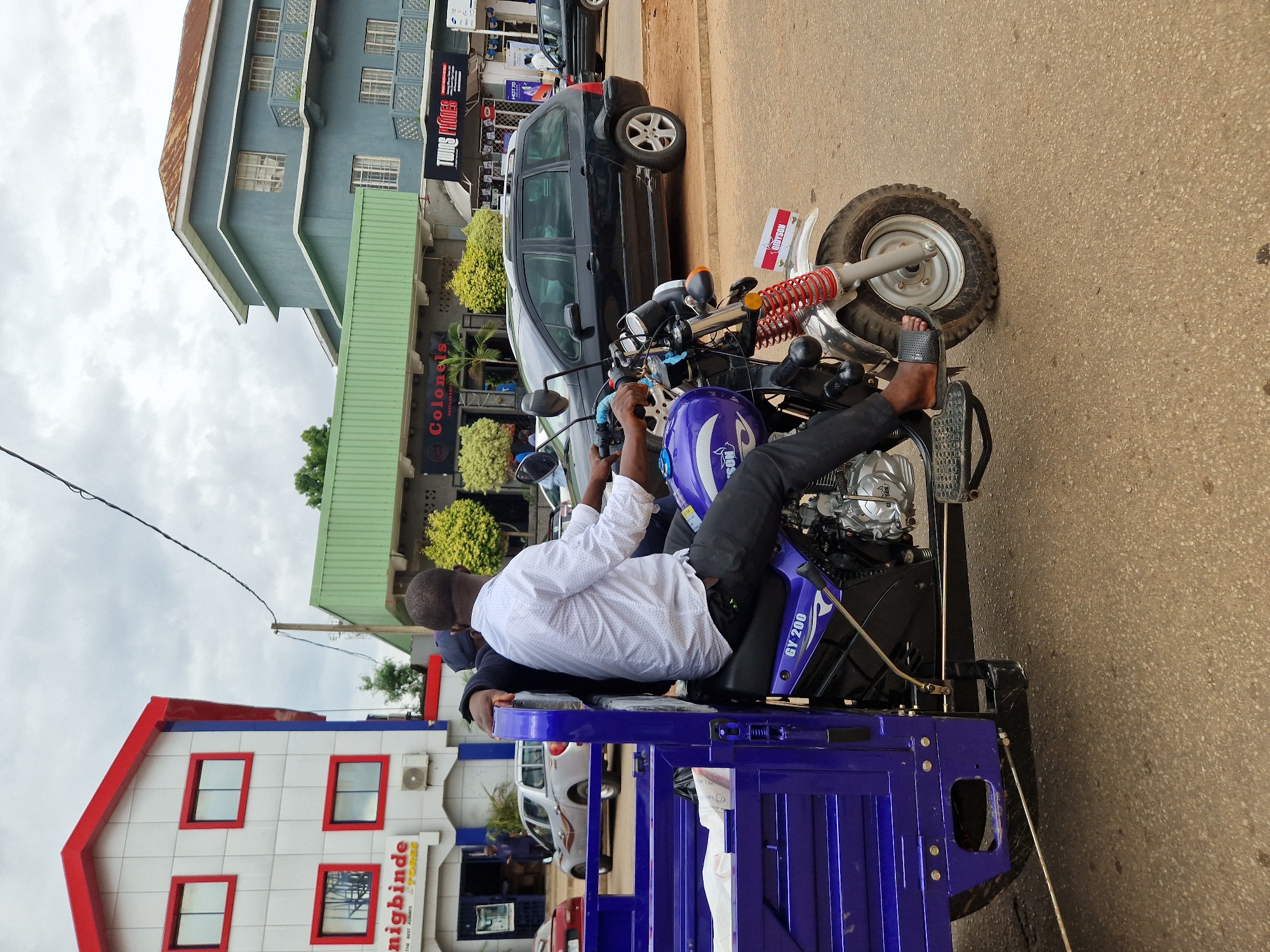 A man is sitting on a blue motorized three-wheeled vehicle. He is wearing a white long-sleeved shirt and dark pants. He is holding the handlebars of the vehicle, and his left foot is resting on a pedal. The vehicle has a large front wheel and two smaller wheels at the back. There is a cargo bed behind the driver's seat, which is also blue. A black car is parked behind the three-wheeled vehicle. In the background, there are buildings and trees. The sky is cloudy, suggesting it might be an overcast day. The scene appears to be a street in a town or city, with shops and commercial buildings visible. The man's posture suggests he might be preparing to drive or has just stopped. There is text visible on the side of the three-wheeled vehicle that reads "GY 200". On a banner attached to the vehicle's fender, text in red and white appears. Additionally, there are signs on the buildings, including one with red lettering that says "Colonels RESTAURANT". Another building has a sign with red and white text that says "nigbinde STORES". The image is oriented vertically, with the top of the image appearing on the left.