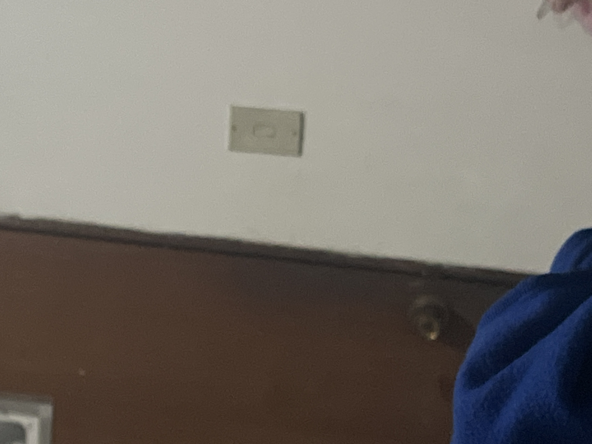 The image depicts a close-up view of a wall with a light switch and a portion of a brown door. A person's shoulder and a bit of their blue garment are visible in the lower left corner. The setting appears to be indoors. There is no indication of the location within the city of Santa Ana, Venezuela.
