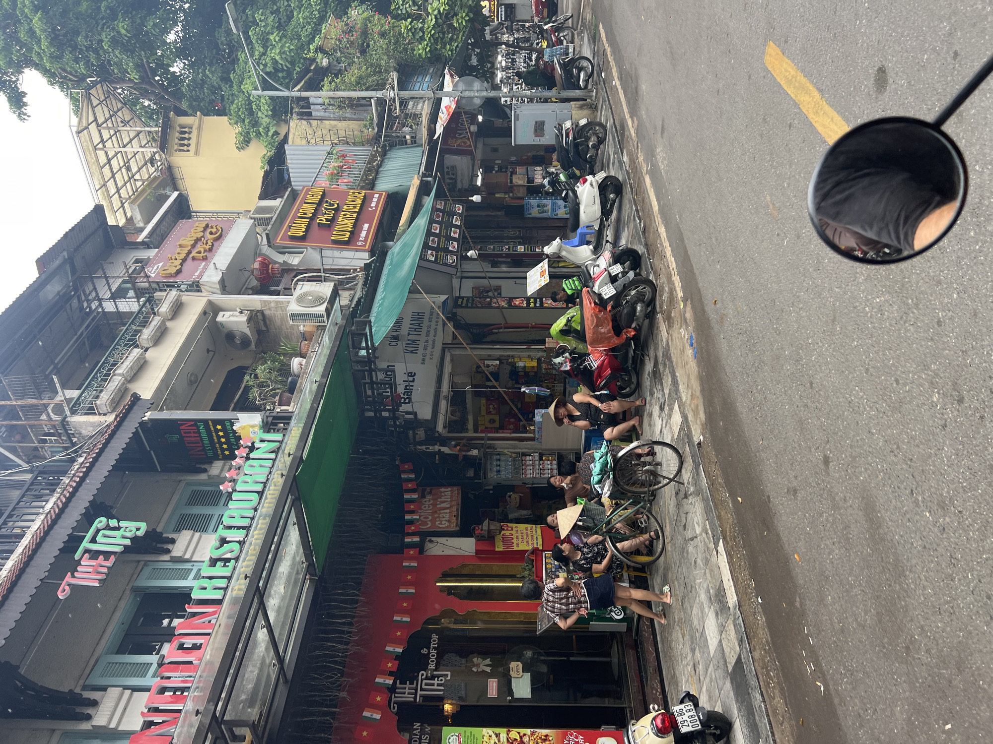 The image depicts a street scene in Hanoi, Vietnam, likely in the Old Quarter, characterized by its narrow buildings with balconies and awnings. In the foreground, a portion of a motorcycle handlebar and rearview mirror is visible, reflecting a person's arm and shoulder wearing a black t-shirt. The street itself is paved and appears wet, with a yellow painted line on the right side.
Along the sidewalk, several motorcycles are parked. A red scooter with the license plate "29-BG 008.83" is on the far left. Further down, a white scooter and several other dark-colored motorcycles are parked in a row.
The buildings lining the street house various businesses. On the left, a restaurant named "The Taj Indian Restaurant & Rooftop" is prominently displayed with colorful signage in English and what appears to be Vietnamese. Indian flags are strung across the facade. Next to it, a shop displays "NƯỚC ÉP" (juice). Further along, a sign for "QUÁN NGON Phở Cổ OLD QUARTER DELICACIES" is visible.
Several people are present on the sidewalk. Three women are seated near a bicycle in front of the restaurant. One woman is wearing a conical hat. Another woman stands nearby, and two other women are seated on a bench or low wall. Further down the street, a person in dark clothing is tending to a motorcycle, and another person is partially visible behind a display of products. The buildings are multi-story with external air conditioning units and balconies. Trees with dense foliage are visible on the right side of the image, suggesting an urban environment with greenery. The sky is overcast.