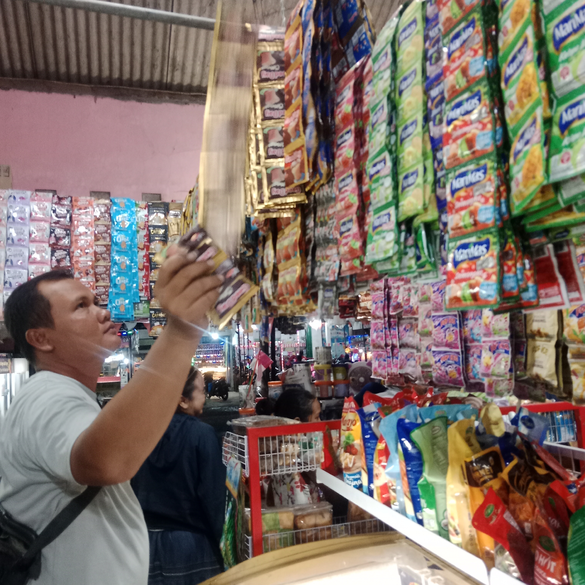 In the city of Tegowanu Wetan, Indonesia, a man is browsing a market stall filled with packaged snacks and drinks. He holds a few items in his hand, examining them. The stall is well-stocked, with rows of colorful packaging hanging from the ceiling and displayed on shelves. Other shoppers are visible in the background, and motorcycles are parked nearby. The market appears to be outdoors, with a corrugated metal roof overhead. The overall impression is one of a bustling local marketplace.