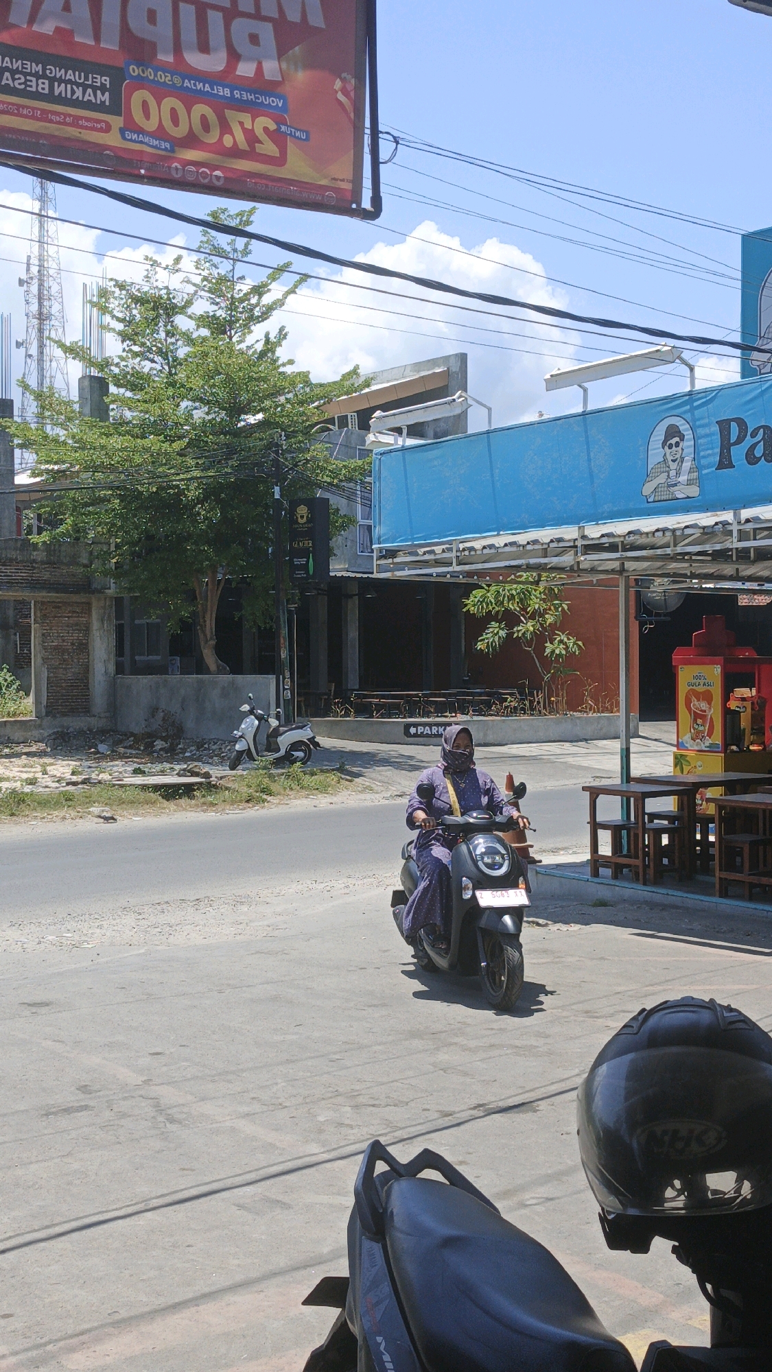 A woman wearing a purple dress and a hijab is riding a black scooter on a paved road in Desa Pagandaran, Indonesia. The scooter has a license plate that reads "B 5663 YY". The woman is looking forward and appears to be alone. The environment is urban, with buildings and shops visible in the background. There are also trees and power lines. The sky is blue with some clouds. In the foreground, the back of another scooter and a black helmet are visible.