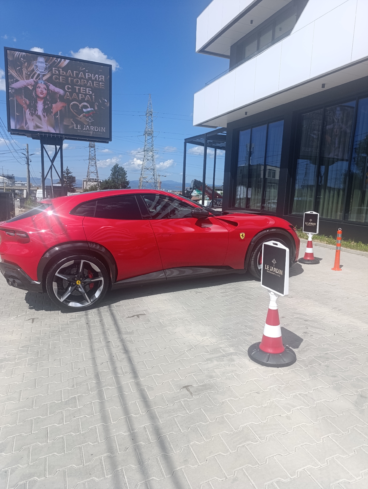 A red Ferrari is parked on a paved area in front of a modern building. The building has large glass windows and a white facade. Two white signs with black lettering that read "LE JARDIN RESTAURANT & GRILL" are placed on traffic cones near the car. In the background, a large billboard displays an advertisement with a woman, a microphone, and a Bulgarian flag. Several power line towers are visible in the distance. The sky is blue with some clouds. The scene appears to be outdoors in a city environment, likely Sofia, Bulgaria, based on the Bulgarian text on the billboard. There are no people in the immediate vicinity of the car, though the billboard suggests a public advertising space.