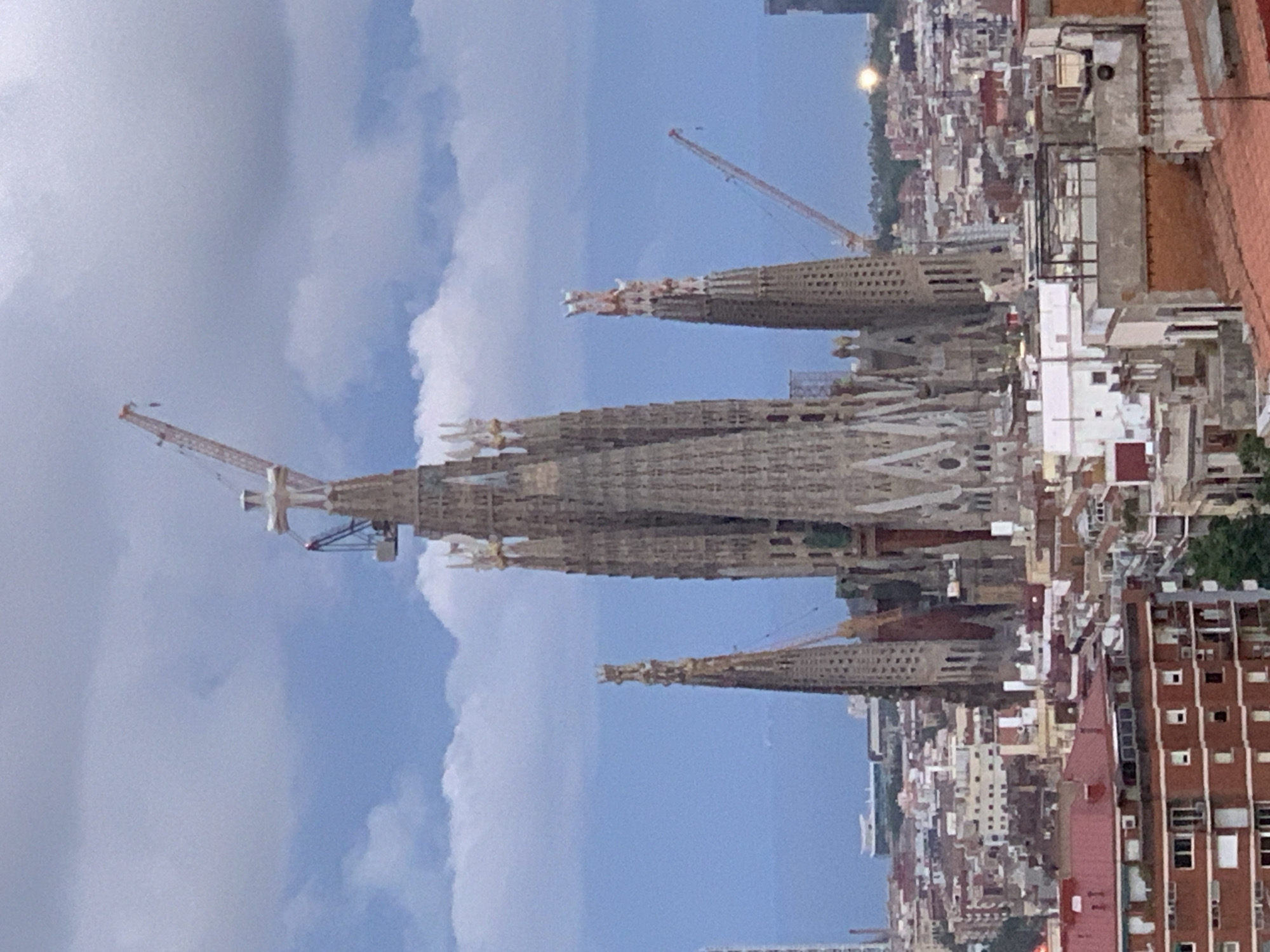The image shows the Sagrada Familia, an unfinished basilica in Barcelona, Spain. The structure is under construction, with cranes visible. The building towers over the surrounding urban landscape, which is densely packed with buildings. The sky is partly cloudy.