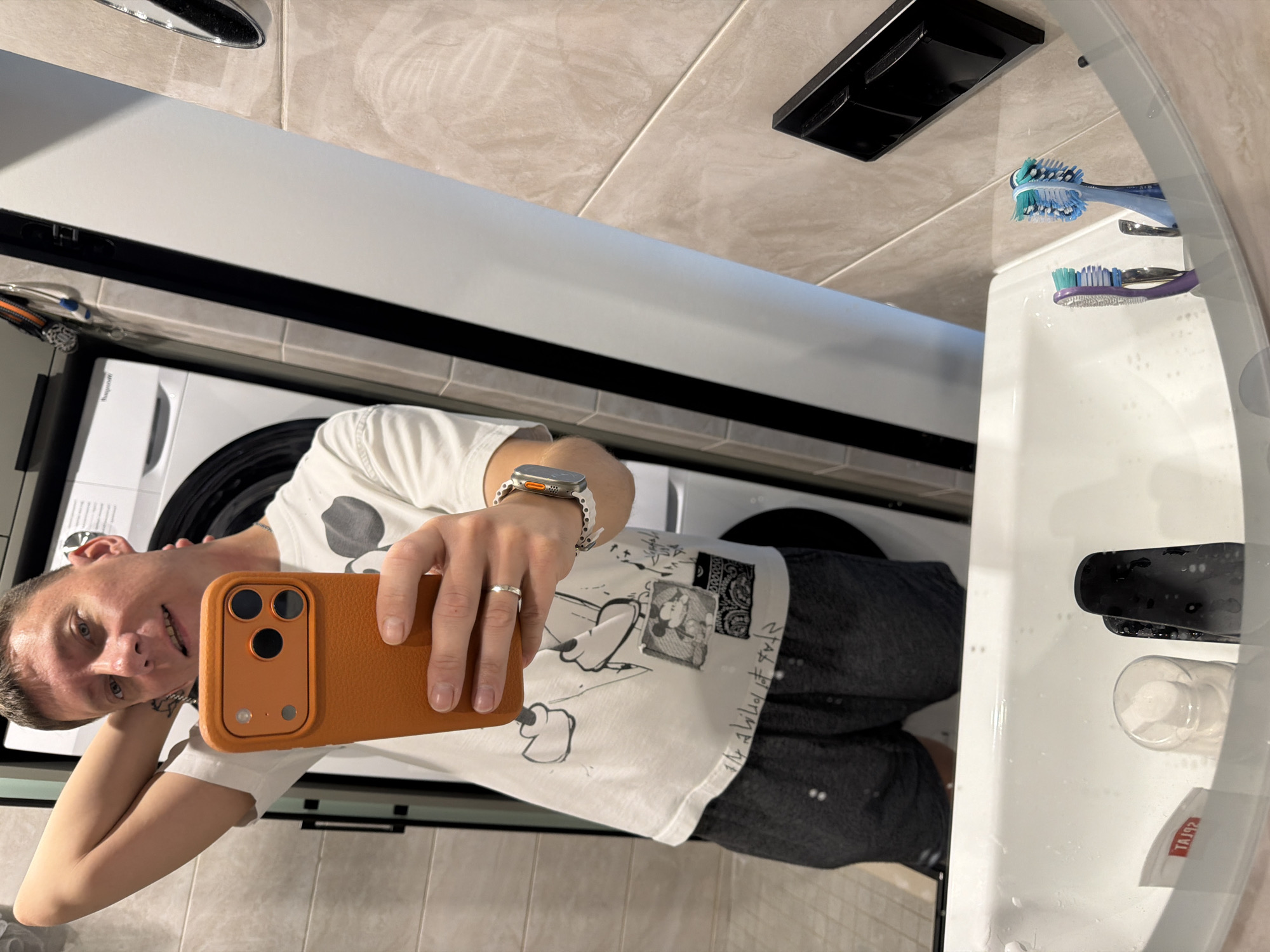 A man takes a selfie in a bathroom. He is wearing a white t-shirt with a Mickey Mouse graphic and dark shorts. He is holding his phone, which has an orange case, in his right hand, and has his left hand behind his head. A washing machine is visible behind him. In the foreground, a white sink is filled with water, and two toothbrushes and a bottle of mouthwash are visible. The scene appears to be in a residential bathroom.