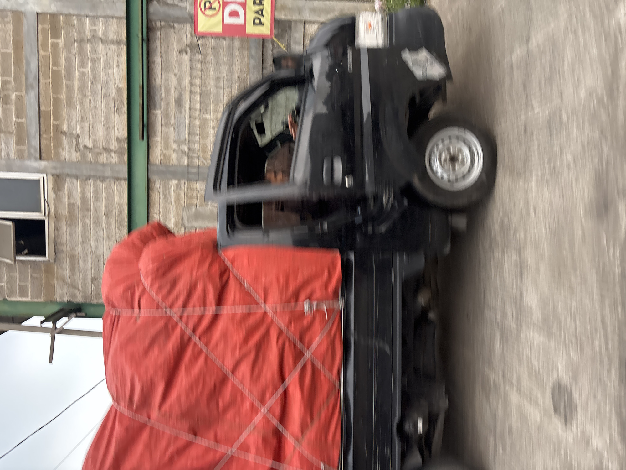 A black pickup truck with a red tarp covering its cargo is driving on a road in Bekasi, Indonesia. The truck has a driver and one passenger visible. The background features a brick building with green metal beams. A yellow sign with red text is also visible on the right side of the image. The scene is captured with motion blur, indicating the vehicle is in motion.