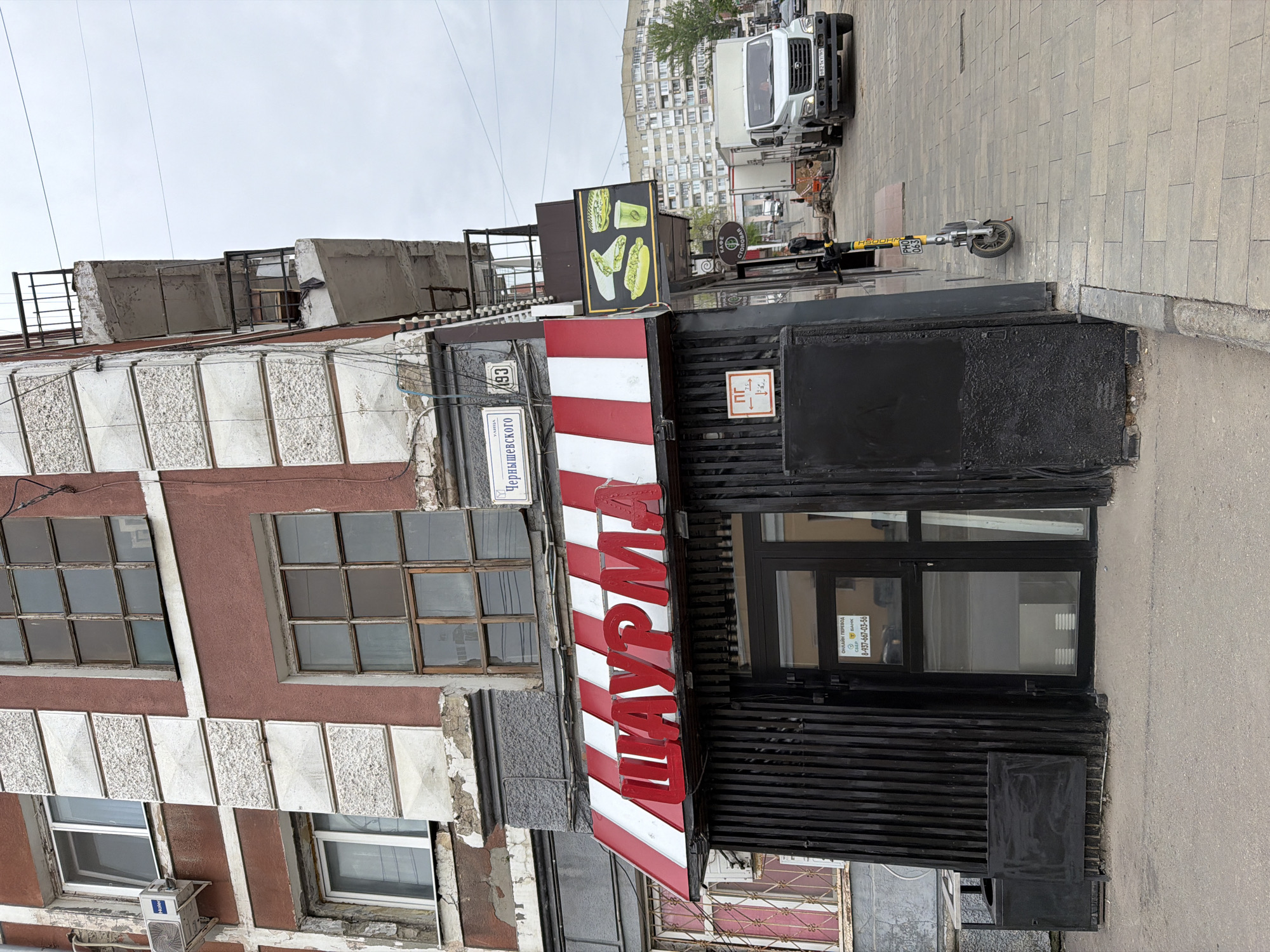 A ground-level shot depicts a storefront in Saratov, Russia, identifiable by the street sign "Чернышевского" (Chernyshevskogo) attached to the building's facade. The sign includes the number "193." The storefront has a red and white striped awning bearing the Cyrillic letters "ШАУРМА" (Shaurma), indicating that the business sells shaurma, a type of wrap or sandwich.
The building adjacent to the store is a multi-story structure with a facade of alternating horizontal rows of white and reddish-brown panels, with a pattern of square windows. An air conditioning unit is visible on the wall.
The storefront itself has a modern, boxy construction with black vertical slats. The door appears to be glass. There is a scooter parked on the sidewalk in front of the store. A white van is parked further down the street. Additional signage advertising various food items is present. The overall setting is an urban environment with other buildings visible in the background.