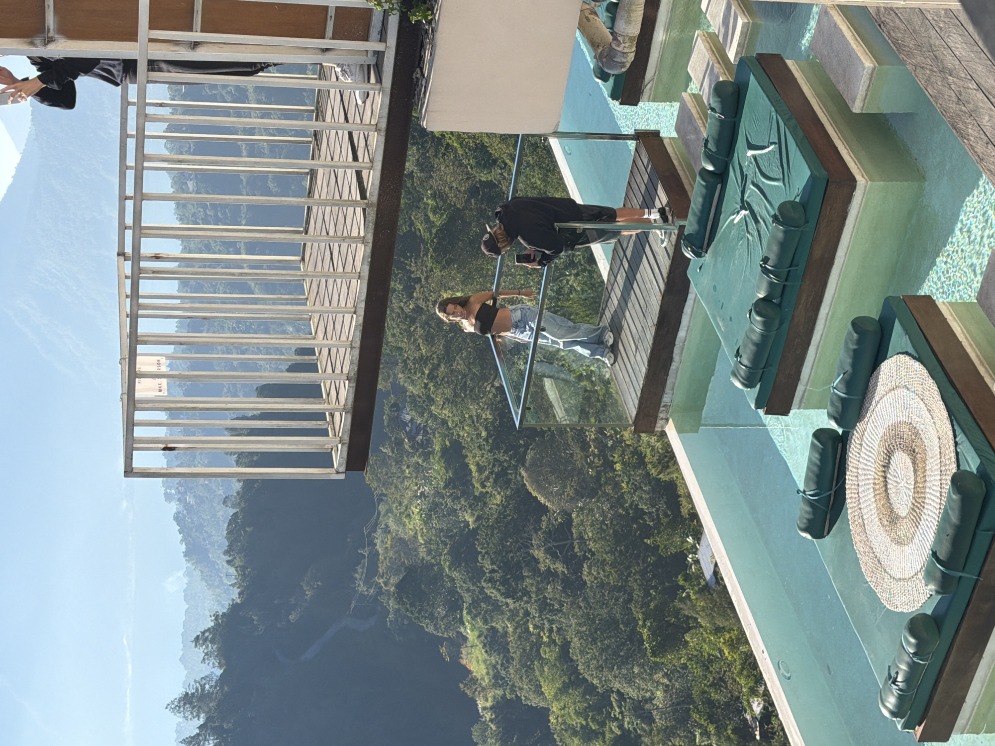 This image depicts a multi-level outdoor resort or viewpoint located in Kintamani, Indonesia, offering extensive views of a forested valley and distant mountains under a clear blue sky.
In the upper section, a person is partially visible on the far right, standing on a metal-railed balcony with vertical bars. This individual wears a black long-sleeved top, dark pants, and white sneakers, and holds a smartphone elevated, seemingly capturing the distant landscape. A partially visible sign on the railing includes the text "MAX PERSON".
Below this, on a wooden deck featuring a glass railing, a woman wearing a black strapless crop top, loose light blue jeans, and white sneakers leans against the railing, looking to her right, appearing to pose. To her right, a man in a black baseball cap, black hoodie, dark shorts, and black socks with white sneakers stands, actively photographing her with a smartphone held in his hands.
Further down and extending from the deck, a multi-tiered infinity swimming pool with light blue water is visible. Within the pool, several floating or submerged wooden platforms are outfitted with dark green upholstered cushions and bolster pillows. The platform closest to the viewer features a large, round woven mat with concentric light brown and white patterns placed on the green cushion. Concrete or stone steps are positioned between some of these platforms.
The background reveals a vast, deep valley covered in dense green tropical forest. A winding road is visible through the foliage in the mid-ground. Hazy blue mountain ranges stretch across the distant horizon, beneath a bright, unclouded sky. The scene is illuminated by strong daylight, casting clear shadows.