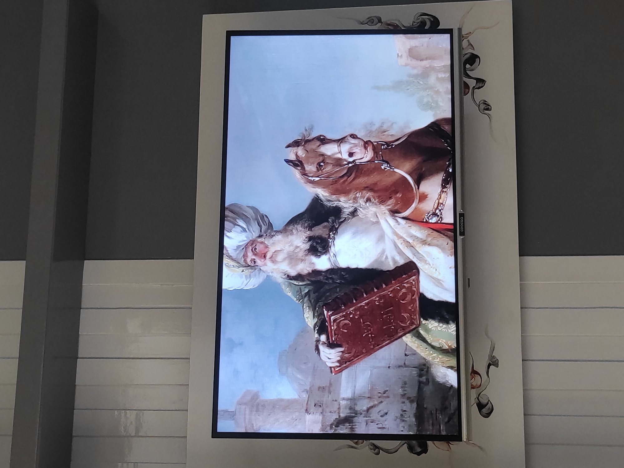 The image shows a television screen displaying a painting of a man with a long white beard and a turban, dressed in ornate robes, seated on a brown horse. He is holding a large, red, intricately decorated book in his left hand. The background of the painting depicts a city with architectural structures and a pale blue sky. The television is mounted on a wall with a decorative frame that includes swirling black and white designs. Below the television, the wall has vertical panels. The text "COOCAA" is visible on the bottom edge of the television. The overall setting appears to be an indoor space, likely a home or a public area in Sumedang, Indonesia, given the context provided.