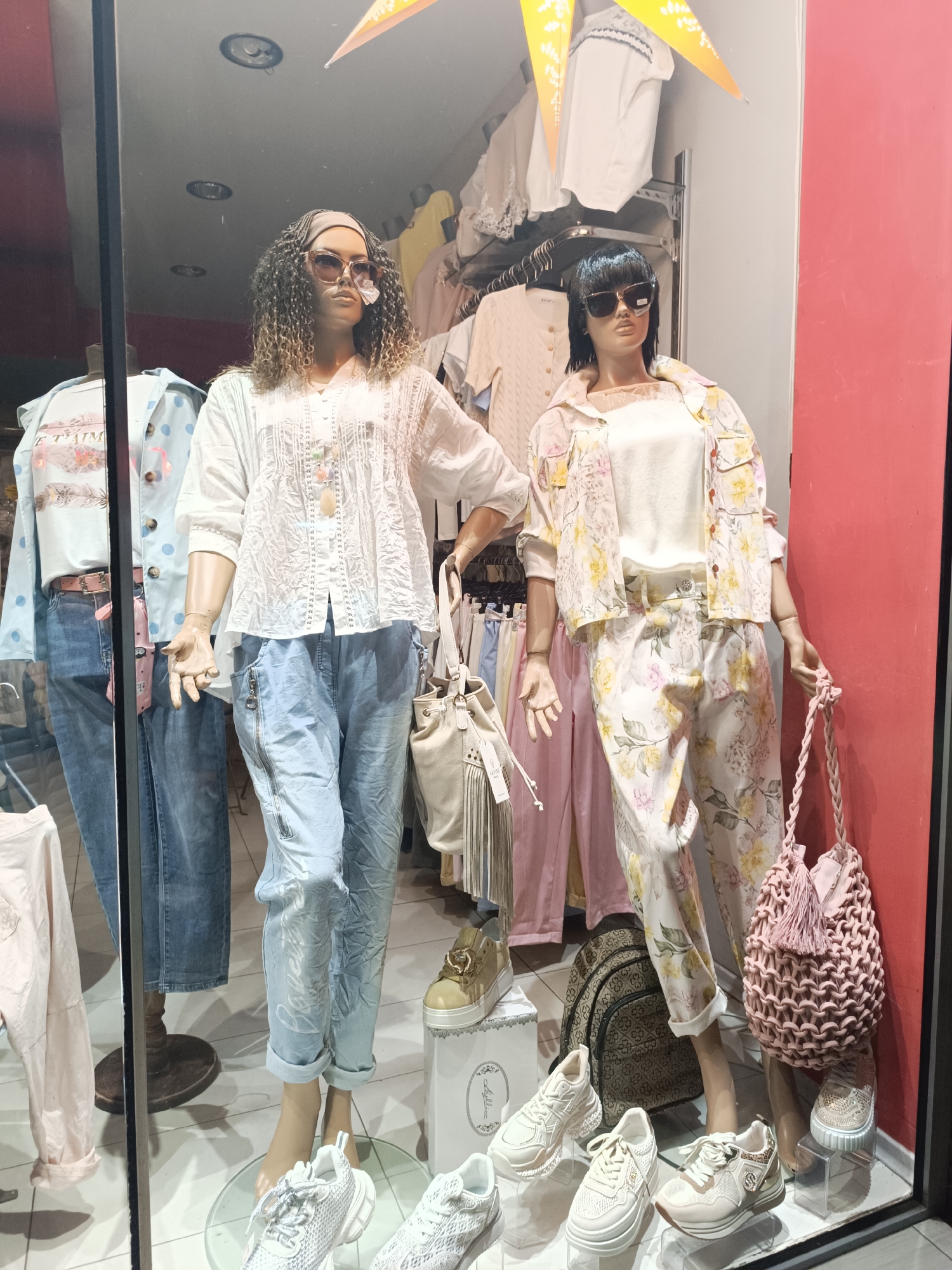 This full-shot image captures a vibrant storefront display of a clothing boutique in Pleven, Bulgaria, showcasing casual spring/summer fashion. Two female mannequins are prominently featured, surrounded by additional clothing and accessories.
The mannequin on the left has curly, ombre-style hair, a headband, and large rectangular sunglasses with a small tag on the lens. She wears a loose, white blouse with intricate lace and embroidered details, paired with light blue, distressed-look capri jeans that feature zippers and subtle white text print. A large, light-colored bucket bag with fringe hangs from her left hand.
The mannequin on the right features short, dark hair and dark rectangular sunglasses. She is dressed in a light-colored camisole under a short-sleeved, collared button-up shirt and matching cuffed pants, all adorned with a cheerful yellow and pink floral pattern. She holds a small, pink knitted or crocheted bag with a tassel.
Both mannequins wear light-colored athletic-style sneakers. The left mannequin's are white with thick soles, while the right mannequin's are beige with gold accents, possibly featuring a 'C' logo.
In the background, clothing racks hold pastel-colored garments. The floor display in front of the mannequins showcases several pairs of trendy sneakers on clear stands, in shades of white, beige, and light pink. A patterned backpack is also visible on the floor. The shop interior's ceiling lights and yellow bunting flags can be seen, along with a red wall on the far right.
Visible text includes "E T'AIME" or "JE T'AIME" on a light blue shirt on the far left, and "Millana" on a white display box. The display effectively presents a relaxed and stylish collection of outfits and accessories.