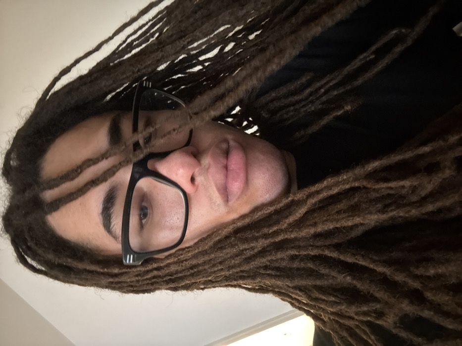A close-up, headshot-style portrait depicts an individual with dark skin, pronounced lips, and long brown dreadlocks that cascade around the face. The person wears thick-rimmed black eyeglasses and a black garment, likely a shirt, visible around the neck. The gaze is directed forward, consistent with a selfie perspective. The background consists of an unidentifiable, plain, light-colored surface, indicating an indoor setting. No specific landmarks or environmental features are present to indicate a location within Paris, France, or any other geographical area.