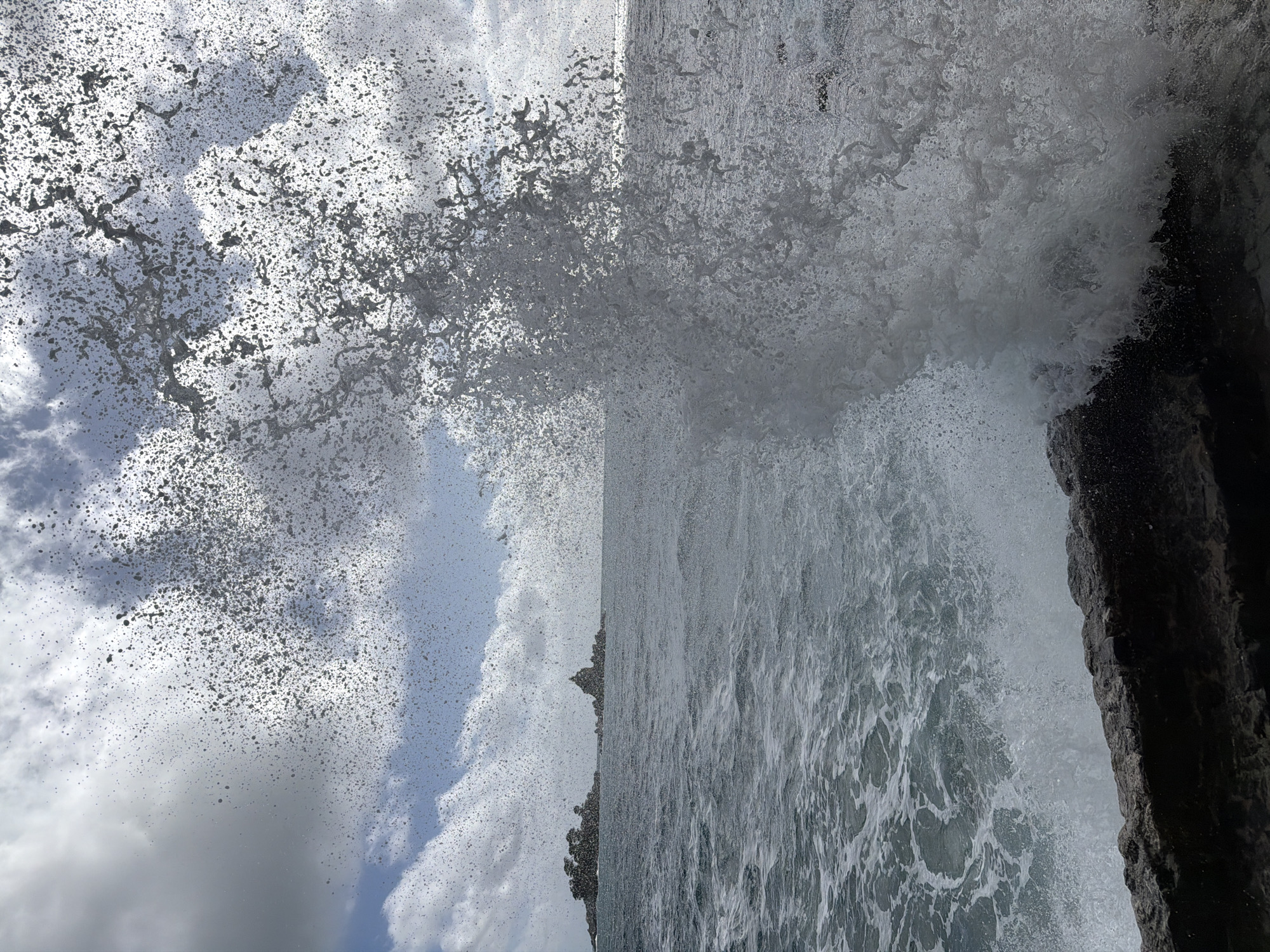 The image shows a large wave crashing against rocks. The location is most likely near Petrovac centar, Montenegro, judging by the coastal characteristics and the prompt's information. Water spray is extensive, reaching high into the air. Sky is visible in the background with clouds. The rock face is dark and rugged. The water is turbulent and foamy. No people are visible.