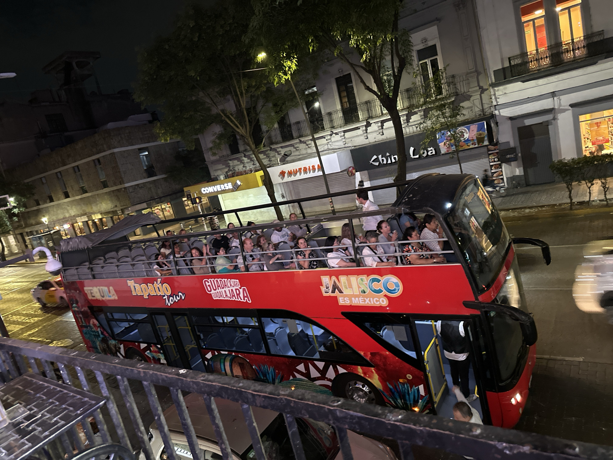 The image depicts a double-decker bus, packed with passengers, in Guadalajara, Mexico. The bus is red and branded with "Tapatio Tour" and "Jalisco Es Mexico." It is traveling on a street lined with buildings, including businesses with signs for "Converse," "Nutrisa," and "Chin Loa." There are trees lining the street, and the scene is lit by streetlights. Pedestrians can be seen near the bus. The photo is taken from an elevated position, behind a metal railing. There is a car parked below the railing.