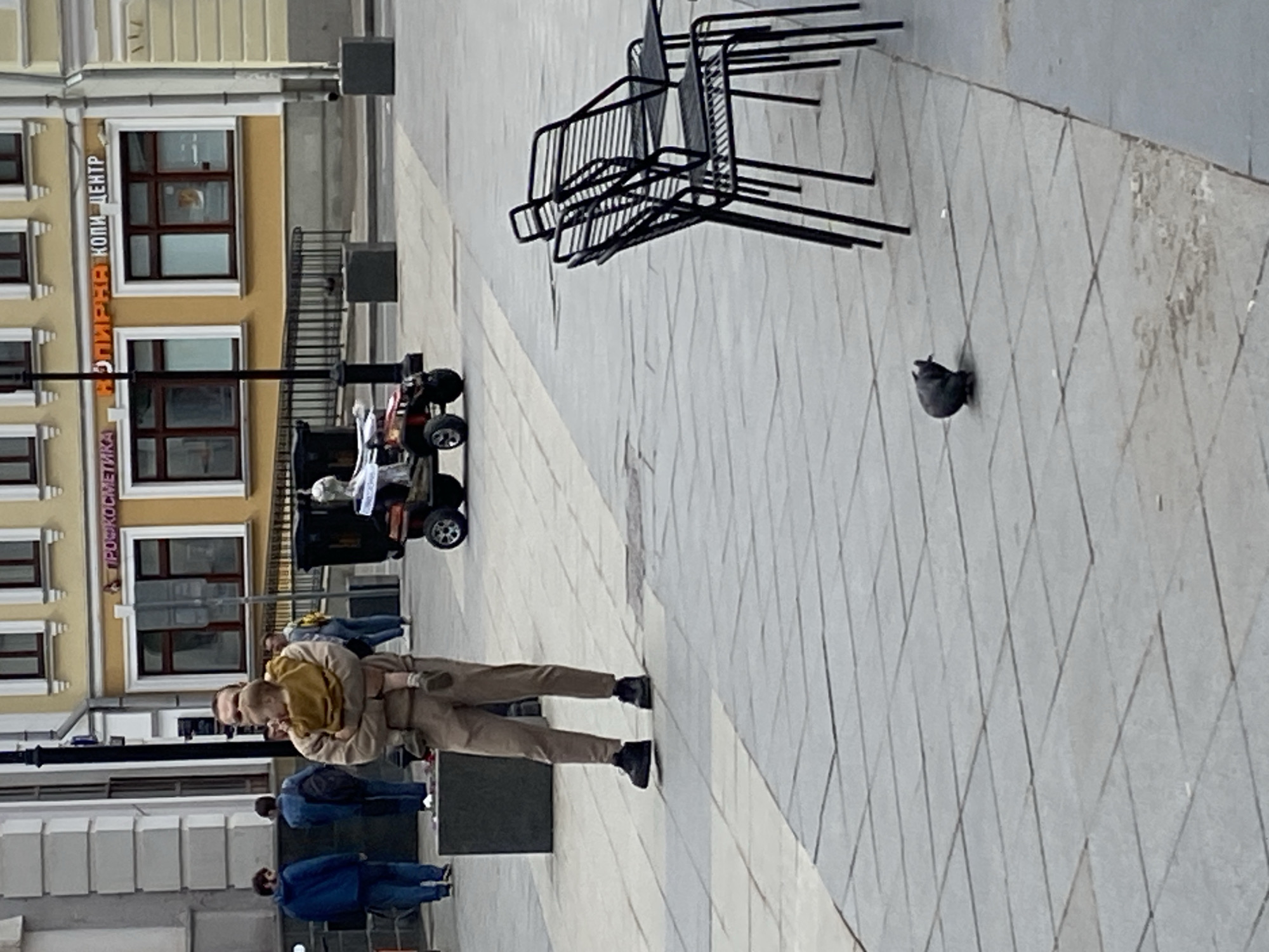 The image depicts an outdoor scene in Moscow, Russia. A man is holding a child. Several people are visible walking in the background. A stack of metal chairs and a small cart are also in view. The ground is tiled. A pigeon is present. Buildings with windows are visible on the left side of the frame.