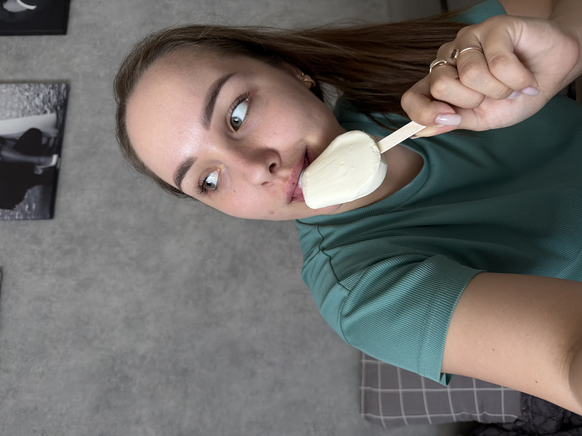 A young woman is holding and biting into a white popsicle. She has brown hair and is wearing a green shirt. She is wearing rings on her fingers. In the background is a gray wall with a black and white picture. A gray and white checkered blanket is also visible. The location is Chaikovsky, Russia.