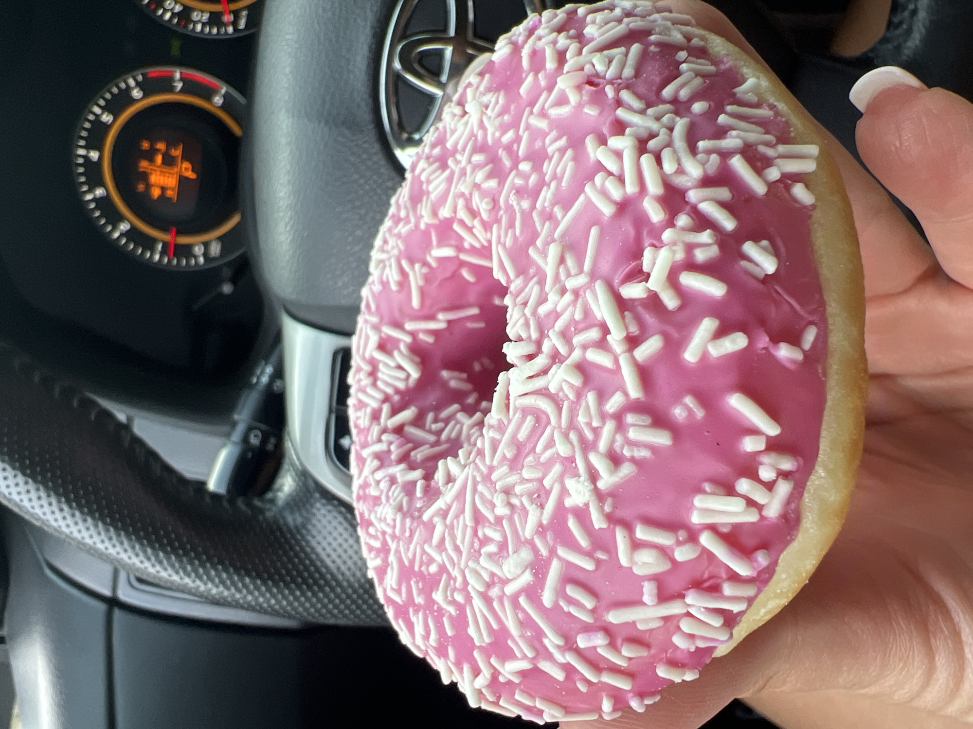 A vibrant close-up shot features a pink-frosted donut covered in white sprinkles, held by a hand inside a car. The donut, brightly colored and in sharp focus, dominates the right side of the frame, showing its soft texture and the slight sheen of the frosting. A human thumb with a white French manicure is visible on the right, supporting the pastry.
The background is a softly blurred car interior. On the left, a segment of a black steering wheel with a textured, possibly carbon fiber-patterned, finish is visible. Behind the steering wheel, a metallic, silver Toyota logo is partially discernible. Further to the left, part of the instrument cluster is visible, displaying an illuminated orange warning light. This light shows a symbol resembling a car outline with a 'P' or a gear graphic, likely indicating a parking brake or transmission status. Numbers like '4', '5', and '6' from a dial are also faintly visible. A black control stalk or lever is seen extending from behind the steering wheel in the upper left background.
The scene captures a casual moment, possibly a snack break, inside a vehicle. The car appears to be stationary, given the illuminated dashboard light. While the image itself doesn't offer specific time-of-day indicators, the overall impression is of an everyday occurrence. The provided context indicates the location as Budva, Montenegro.