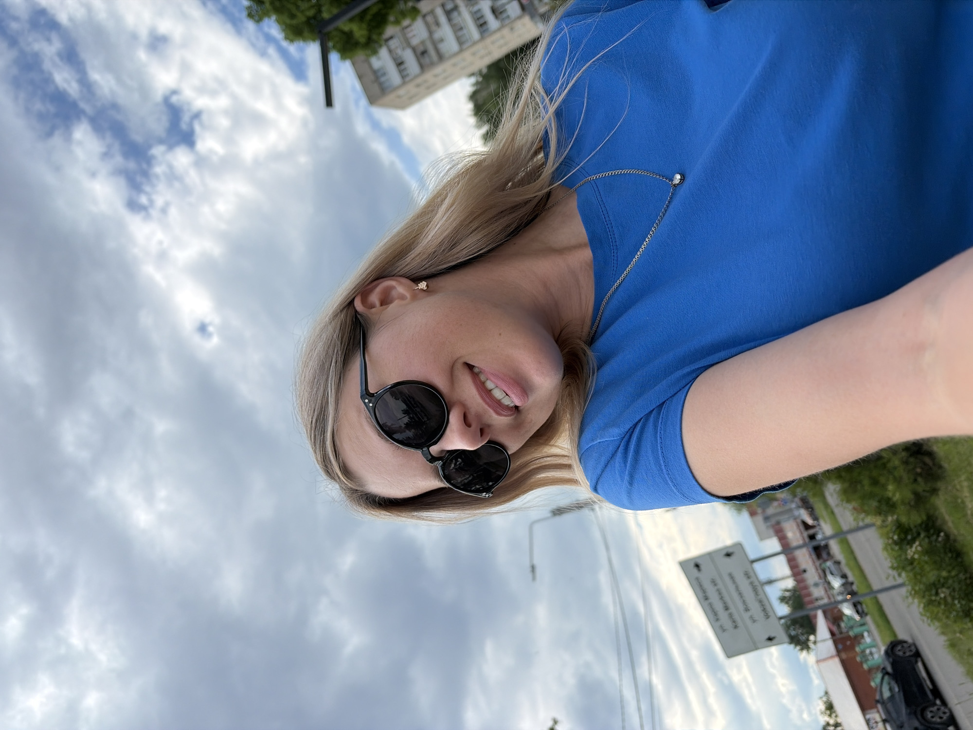 A young woman with blonde hair and sunglasses smiles at the camera outdoors. She is wearing a blue t-shirt and a silver necklace. In the background, there are buildings, trees, and a street sign pointing to "Ul. Yarkaya Marsh", "Ul. Beregovaya", and "Vokzal'naya St.". The sky is cloudy.