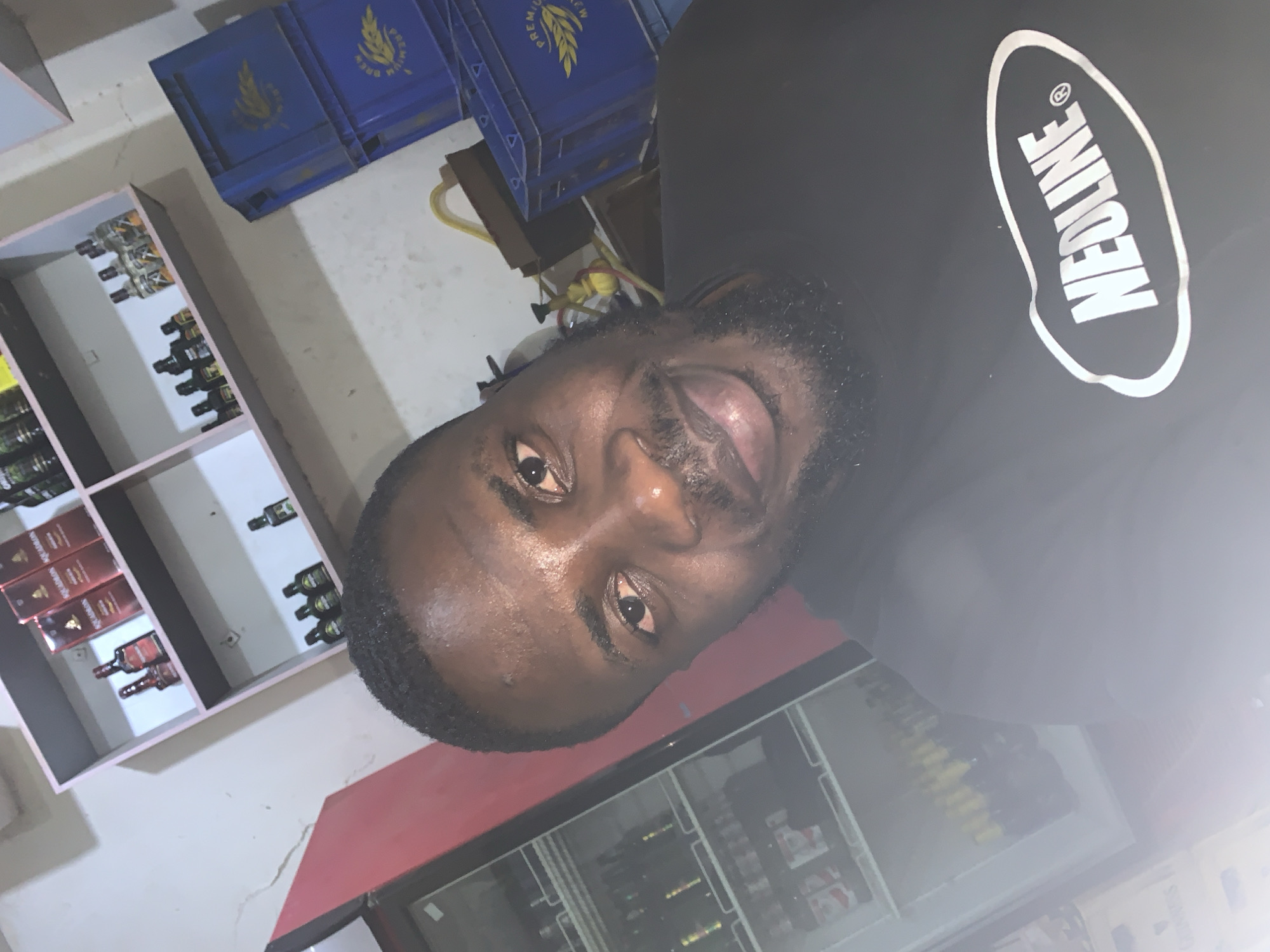 The image is a selfie taken by a dark-skinned man with short black hair and a beard. He is wearing a black t-shirt with a white oval logo that reads "NEOLINE". He is smiling and looking at the camera. The background shows a shelf with various bottles, some of which appear to be alcoholic beverages. There are also blue crates stacked behind him. To the left, a red refrigerator with glass doors is visible, showing shelves stocked with bottles. The setting appears to be a shop or a bar. The lighting is dim, and there is a lens flare in the lower right corner. The man is positioned in the foreground, with his face taking up a significant portion of the frame. The overall impression is of a casual, self-taken photograph in what seems to be a commercial establishment. The location is not specified in the image itself, and therefore cannot be determined to be New Stadium, Nigeria.
