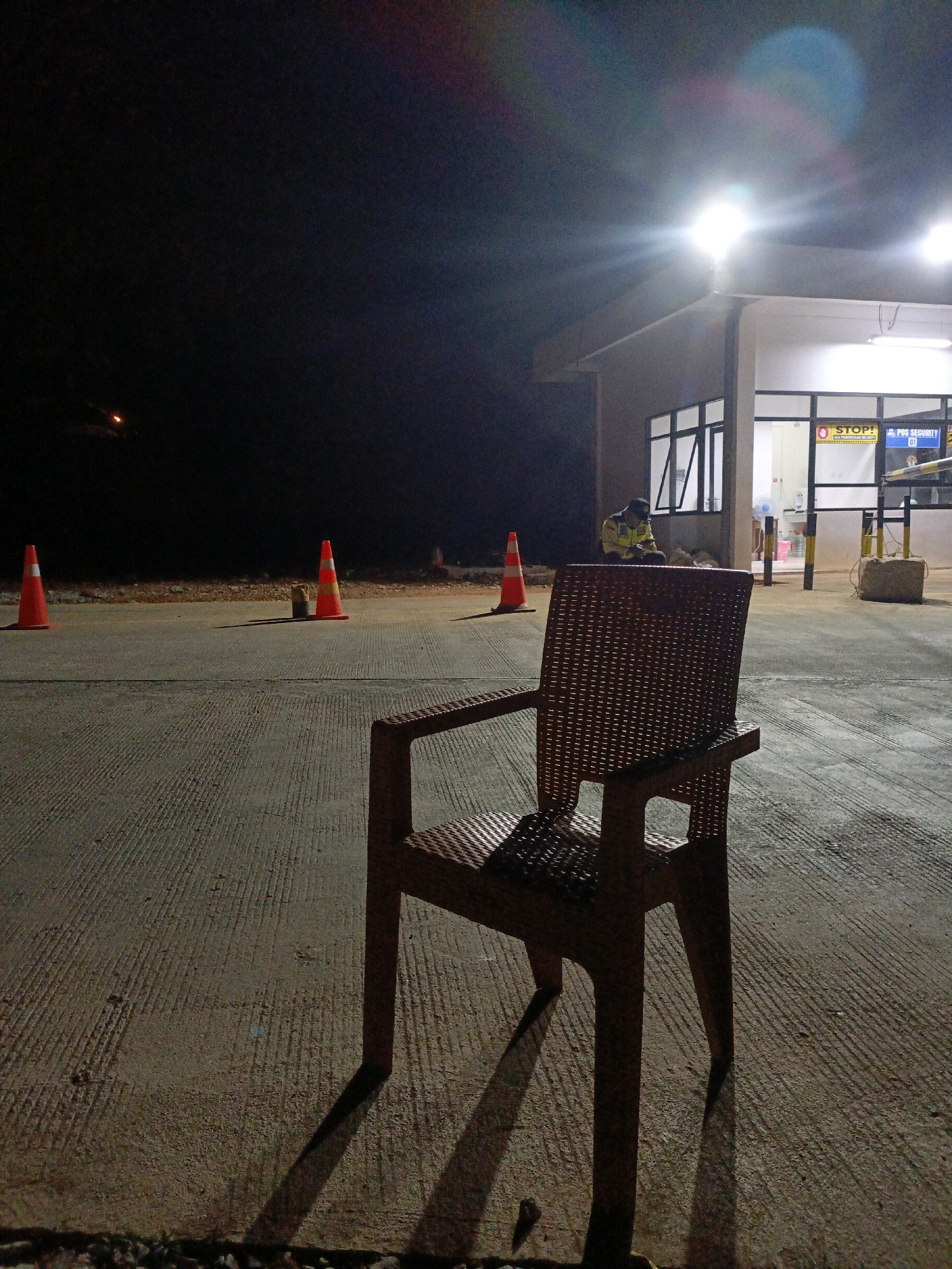 The image depicts an outdoor scene at night, likely in a facility or compound in Jenu, Indonesia. In the foreground, a solitary, dark red plastic chair sits on a textured concrete surface, casting a long shadow to its left. The concrete is marked with parallel grooves. In the mid-ground, several orange traffic cones are placed on the concrete. Behind the cones, a low building with windows is illuminated by bright overhead lights, creating a stark contrast with the dark sky. A person wearing a yellow high-visibility vest and a dark cap is seated near the building, looking down. The building appears to be an administrative or security post, as indicated by signage including "STOP!" and "POS SECURITY 01". The overall atmosphere is one of stillness and quiet, typical of a late-night or early-morning shift.