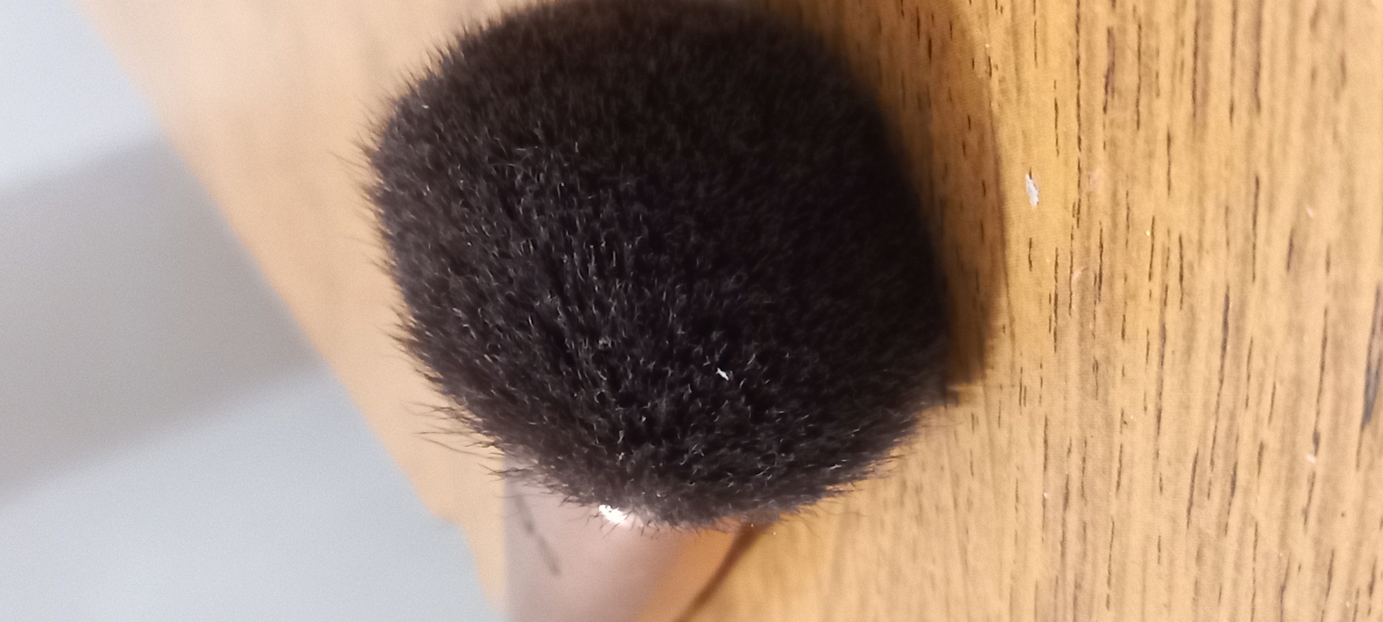 The image displays a close-up of a fluffy, black, spherical object, which appears to be the head of a brush, possibly a makeup brush, resting on a surface. A light brown or wooden handle is partially visible beneath the brush head. The brush's bristles are dense and appear soft, with a few tiny, light-colored particles scattered among them.
The background is divided diagonally into two distinct surfaces. To the right, there is a light brown, wood-patterned surface with visible vertical grain lines and some small, white specks, possibly dust or debris. To the left, the surface is a solid, lighter shade, appearing off-white or light gray, with a subtle shadow falling across it.
The lighting suggests an indoor setting. No specific activity is depicted, as the object is stationary. There is no visible text. The provided context indicates the photo was taken in Kryvyi Rih, Ukraine, though no specific location markers are present in the image itself.