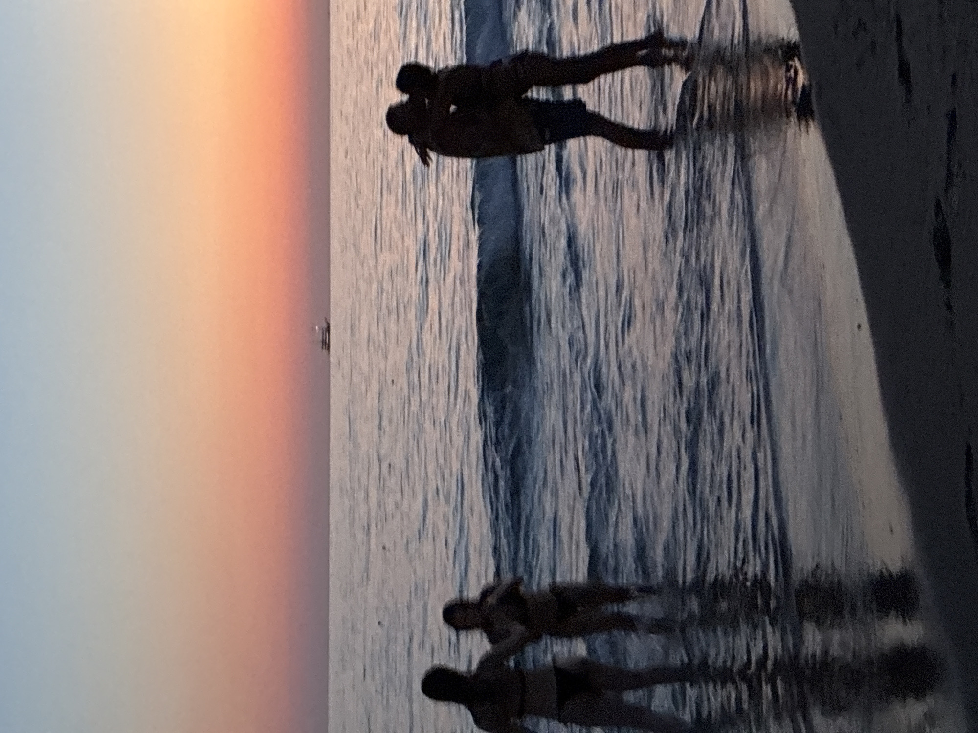 At dusk, four figures are visible in the shallow water of a beach, possibly located in Alanya, Turkey. Two figures on the left, appearing to be female, stand side-by-side. On the right, two figures, one appearing male and the other female, embrace and kiss. The sky displays a gradient of sunset colors, transitioning from pale blue at the top to pink and orange near the horizon. A small boat is discernible on the water in the distance. The waves gently lap at the shore, reflecting the colors of the sky. The scene is captured from a low angle, emphasizing the silhouettes of the individuals against the sea and sky.
