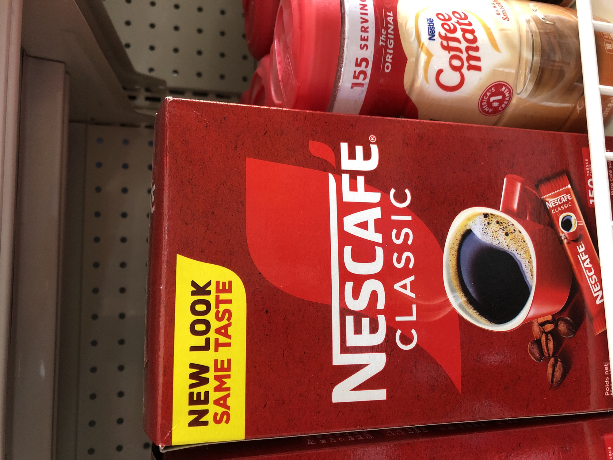 The image shows a close-up view of Nescafé Classic coffee products on a shelf, likely in a retail store. The primary focus is on a red Nescafé Classic cardboard box, which features a yellow banner with the text "NEW LOOK SAME TASTE". Below this, the Nescafé logo and "CLASSIC" are prominently displayed. To the right of the Nescafé box is a container of Coffee Mate creamer, also featuring the Nestle logo. The background shows a white shelf with a perforated texture, and part of another product is visible to the left. The lighting is even, and the image is sharp, suggesting it was taken with a camera or smartphone. There are no people or animals present in the image. The scene is indoors, consistent with a grocery store or supermarket setting. The location of Bar Jirgi Summa, Nigeria, is not discernible from the image itself.