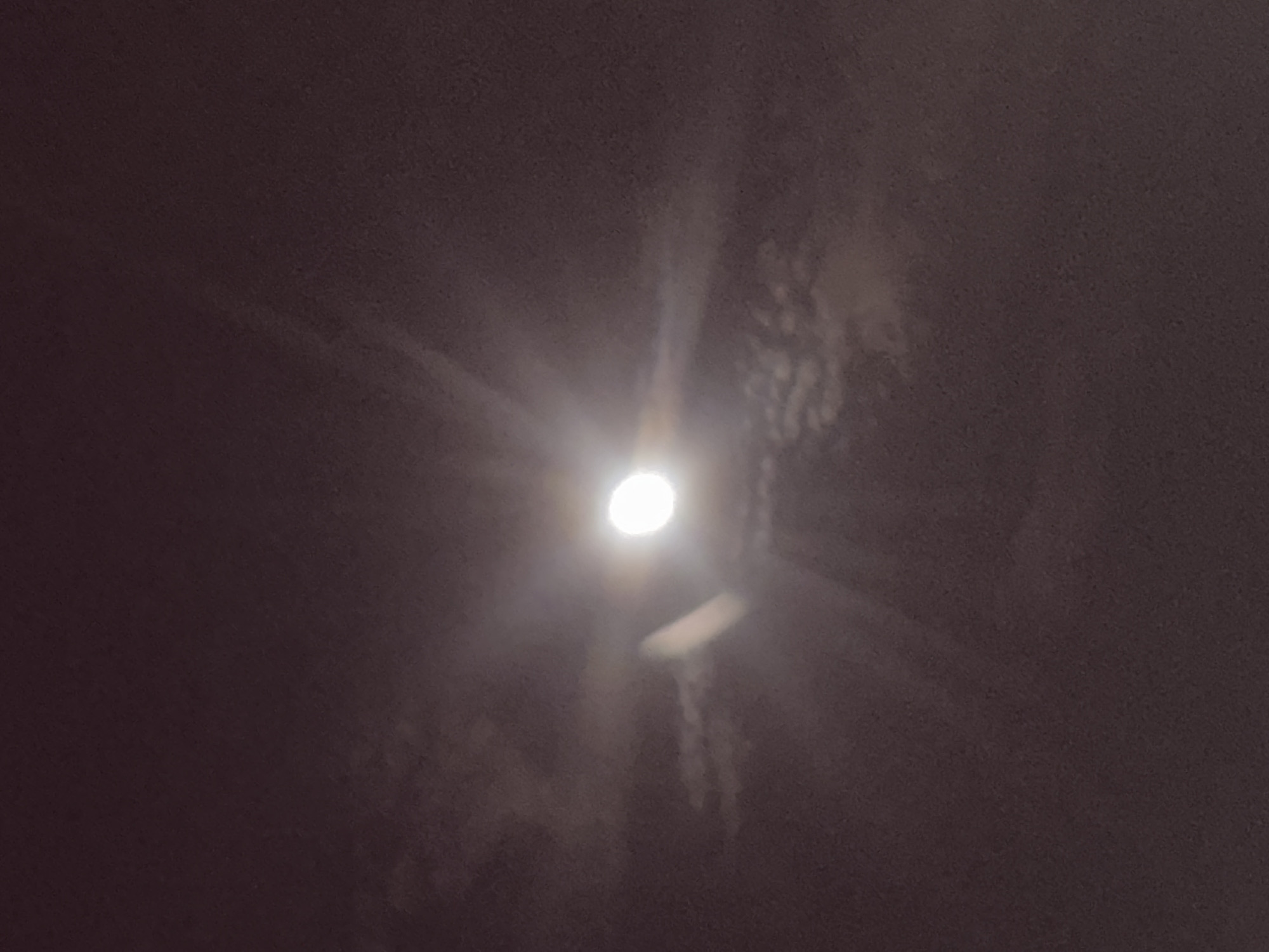 This media file depicts a celestial scene, likely a night sky, with a prominent, bright, full moon as the central subject. The moon is luminous, appearing slightly overexposed due to its brightness, and is partially obscured by faint, wispy clouds or atmospheric haze that create a soft glow around it. Streaks of light radiate outwards from the moon, suggesting its intense illumination.
The surrounding environment is dark and indistinct, with a deep purple-brown hue that indicates nighttime. There are suggestions of indistinct shapes or structures in the background, possibly trees or buildings silhouetted against the night sky, or perhaps more clouds. A long, narrow, light-colored object is visible below and to the right of the moon, possibly a distant plane's contrail, or another celestial object, though its nature is not clear. The overall impression is one of a clear but hazy night, with the moon dominating the view. No people or specific objects are clearly discernible. The time of day is clearly night. The weather is likely clear but with some atmospheric haze or light cloud cover. No location cues are visible in the image itself. There is no visible text.