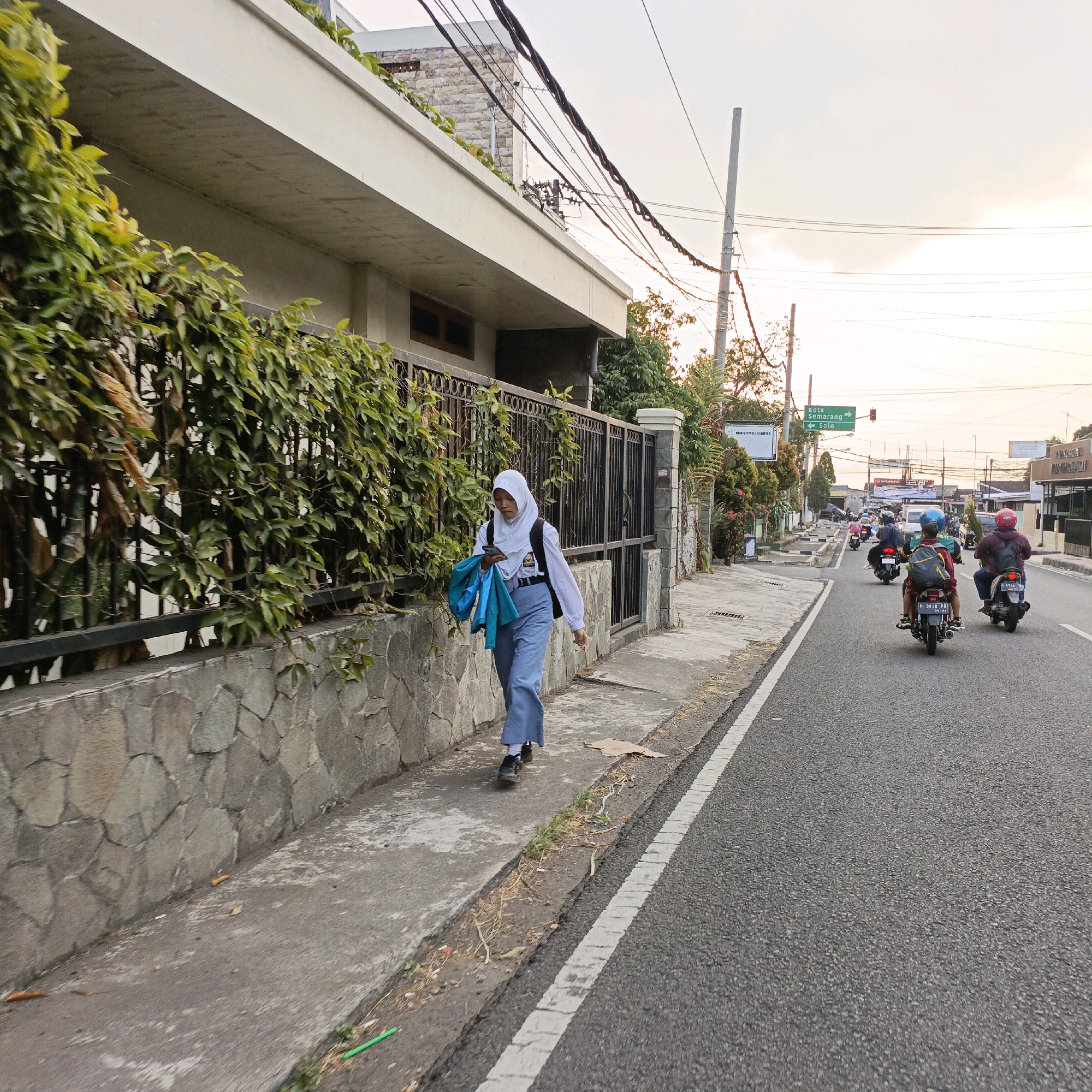 The image depicts a street scene in Salatiga, Indonesia, at what appears to be either early morning or late afternoon due to the warm, soft lighting and the long shadows. On the left side of the frame, a young woman wearing a hijab, a white long-sleeved shirt, and blue pants walks on a sidewalk. She carries a blue bag over her shoulder and holds a mobile phone in her right hand. The sidewalk is bordered by a stone wall and a dense hedge.
The street itself is paved and has a white line marking the edge of the sidewalk. Several motorcycles are traveling on the road, with riders wearing helmets. In the background, there are buildings, power lines, street signs, and vegetation. One sign indicates directions to "Kota Semarang" and "Solo." The overall impression is of a typical urban environment in Indonesia during daylight hours, with ongoing daily activities.
