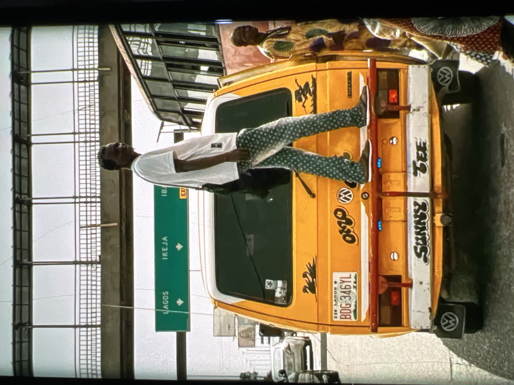The image depicts a street scene in Lagos, Nigeria, likely on a busy road with an overhead bridge. A man wearing a white t-shirt and patterned trousers stands on the back of a yellow Volkswagen van. The van has a license plate that reads "BDG 346YL" and the word "OMO" in a stylized font on its rear. To the right of the van, another man in traditional attire is visible. In the background, a large bus and other vehicles are present. A green road sign indicates directions to "LAGOS," "IKEJA," and "IB." The overall impression is one of urban transit and daily life in a bustling city.