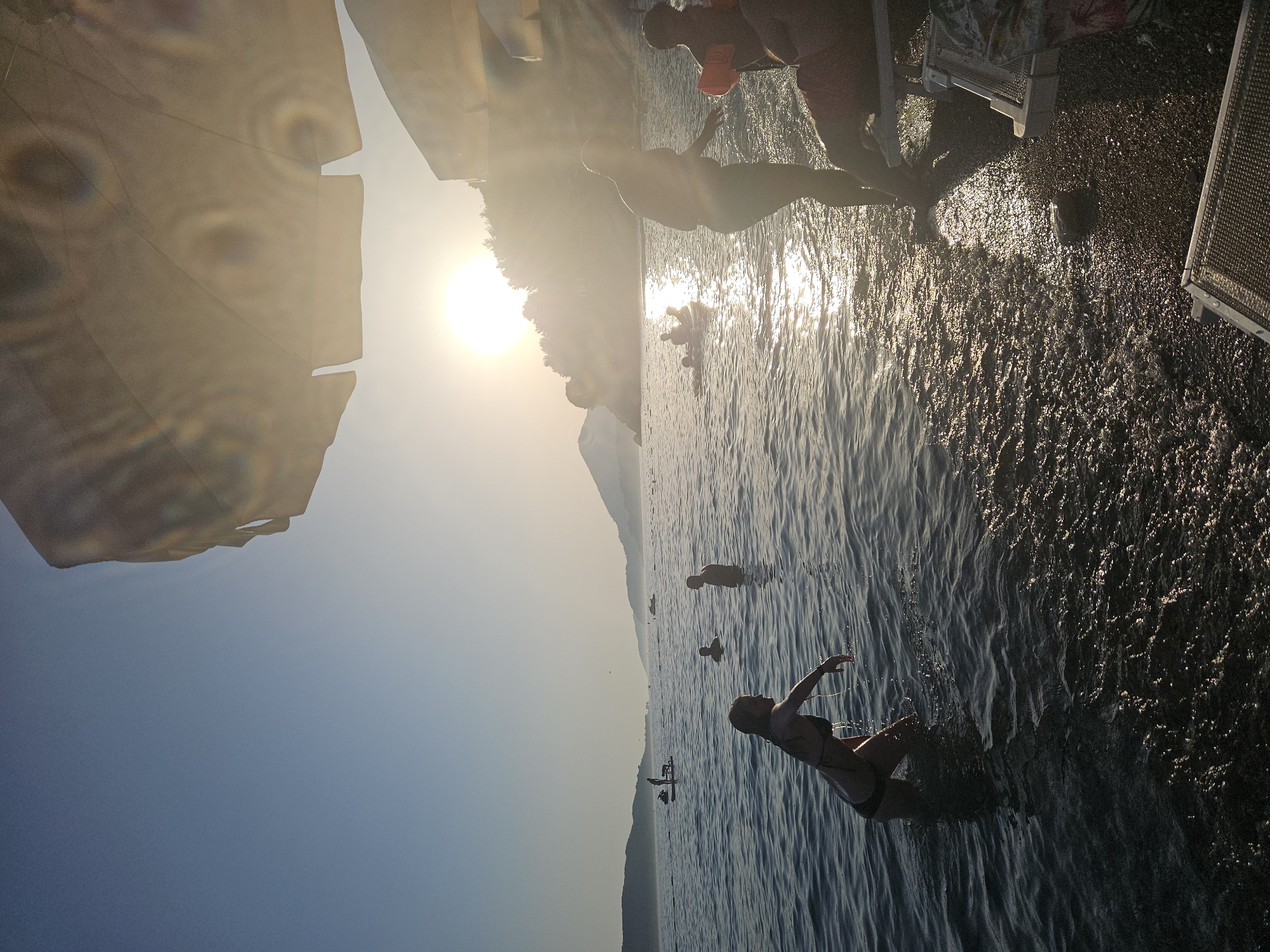 The photograph captures a serene beach scene at sunset. The sun, a bright orb, casts a warm glow over the water, creating a shimmering effect on the surface. In the foreground, a woman in a bikini stands in the shallow water, her left hand reaching out. Further out in the sea, several people are visible, some swimming and others on a small inflatable raft. The coastline is lined with lush green trees and hills in the background, silhouetted against the fading light. Two beach chairs are placed on the shore in the lower right corner of the image, suggesting a relaxing atmosphere. The overall mood is tranquil and idyllic, evoking a sense of peace and relaxation.