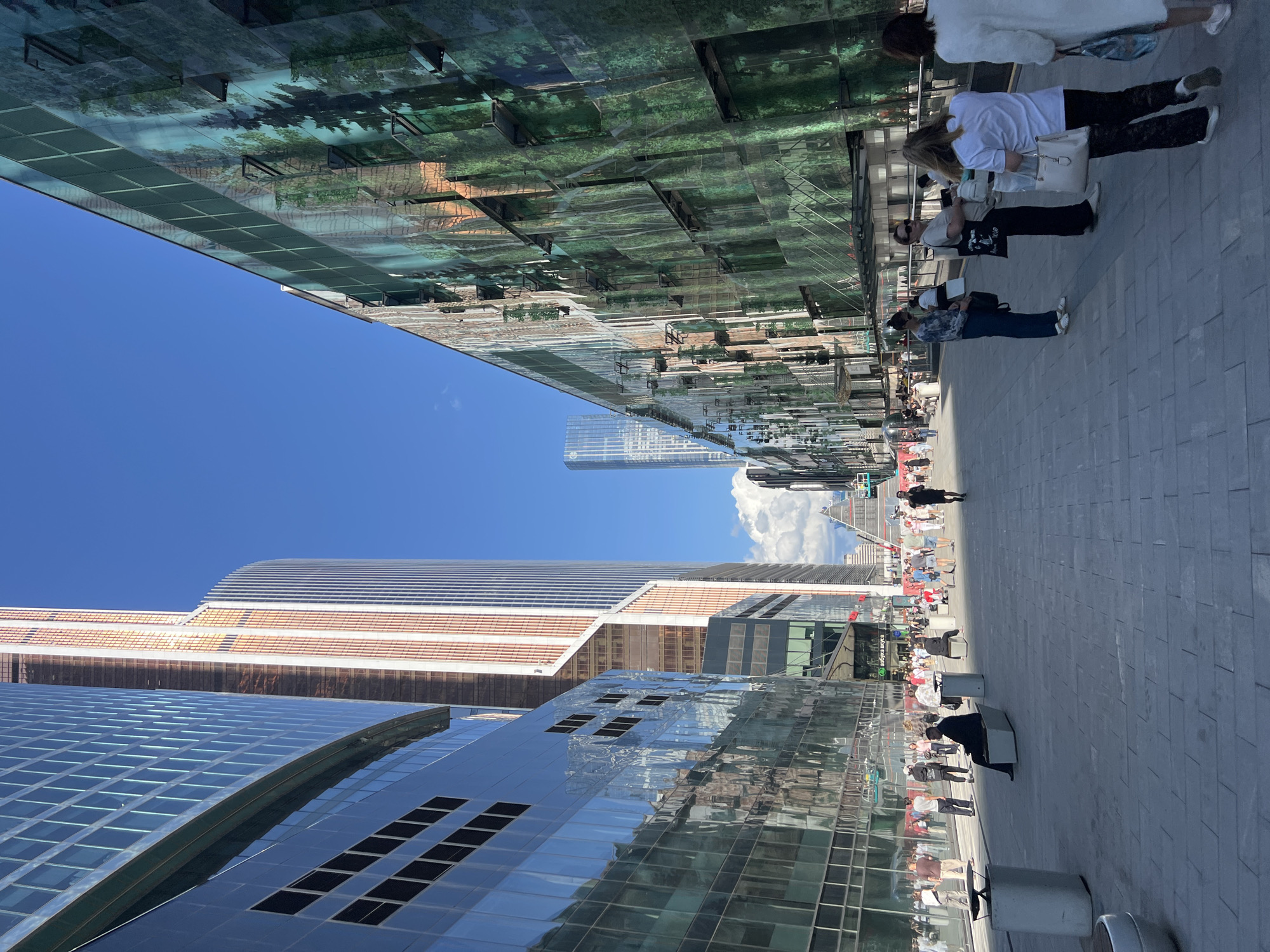 The image depicts a modern urban street scene, likely in Moscow, Russia, due to the architectural style and signage visible. Tall, sleek skyscrapers with reflective glass facades dominate the left side of the frame, interspersed with buildings featuring abstract, forest-like murals on their exteriors on the right. The sky is a clear, bright blue with a few scattered clouds.
A wide, paved pedestrian walkway stretches through the center of the image, populated by numerous individuals. People are seen walking in various directions, some casually strolling, others appearing to be in transit. Several individuals are paused on the walkway, with some seated on modern, angular benches.
On the left side of the walkway, a man in a dark suit is seated on a bench. Further down, a woman in a white top and black pants is engaged in conversation with another woman. To their right, a group of women are standing and chatting. A man wearing a white shirt and dark pants is walking away from the viewer.
In the background, more people are visible, some gathered in small groups. A prominent sign with Cyrillic script, possibly indicating a store or establishment, is visible on a building in the middle ground. In the far distance, several more skyscrapers pierce the skyline, including one with a distinctive, multi-faceted design. The overall impression is one of a bustling, contemporary urban environment on a sunny day.