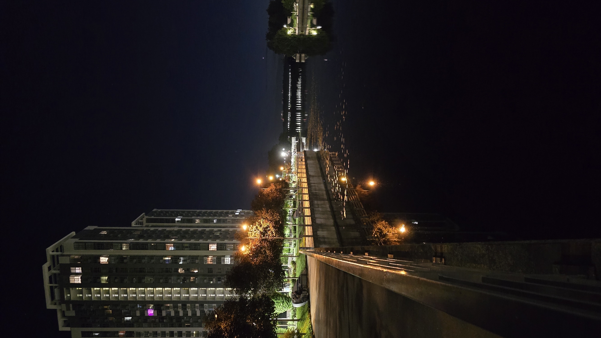 This is a vertical, full-shot, night-time photograph taken at the Obolonska Naberezhna (Obolon Embankment) in Kyiv, Ukraine. The foreground features a concrete embankment with a metal railing, illuminated by streetlights. To the left, a modern, tall residential building with many lit windows is visible. Trees with illuminated leaves line the embankment, and further in the distance, a bridge is visible spanning across the Dnipro River. The river itself is dark, reflecting the lights from the embankment and the distant city. The sky is a deep, clear blue, with a few faint stars visible. There are no prominent human figures in the image, though there appear to be two people sitting on a bench in the middle ground, near the trees. The overall scene depicts a peaceful, urban nightscape along the river.