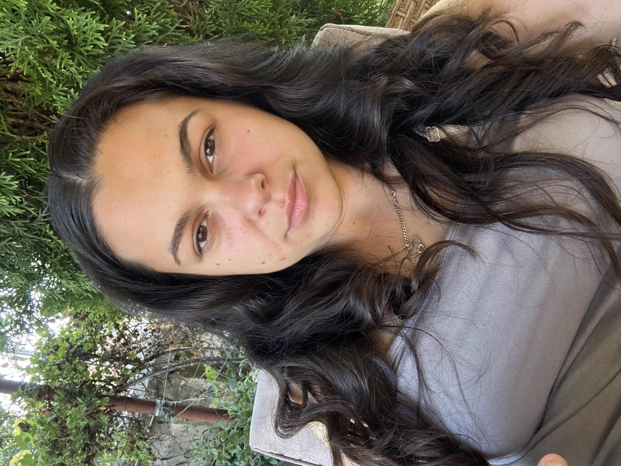 A young woman with long, dark, wavy hair and brown eyes is seated outdoors, possibly on a patio chair. She is wearing a grey tank top and a silver chain necklace with a heart pendant. Behind her, there is lush green foliage, including trees and bushes, suggesting a garden or park setting. The lighting indicates it is daytime, and the overall scene appears relaxed and casual. The location is specified as Oreshak, Bulgaria.