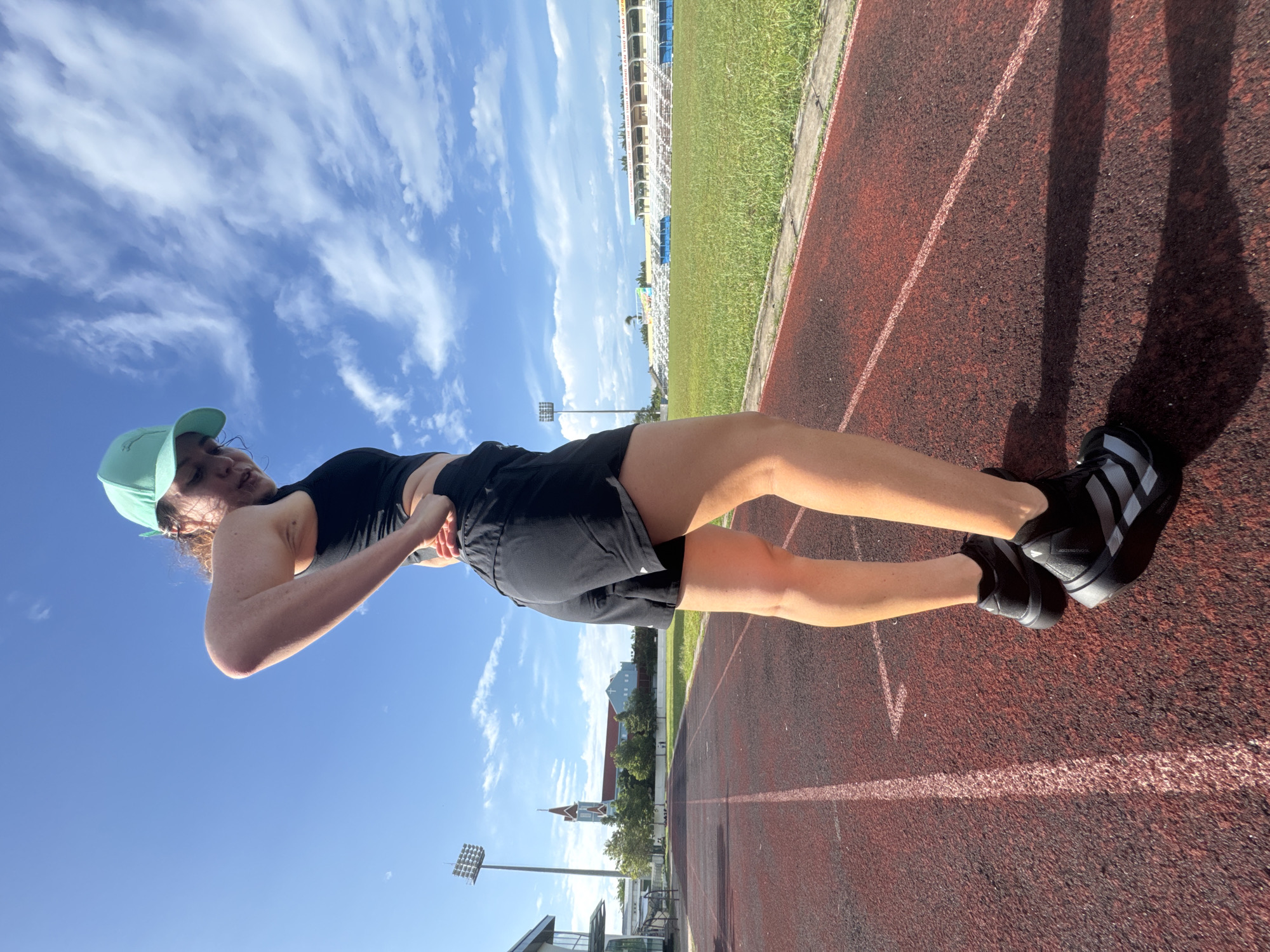 An athletic young woman wearing a black crop top, black shorts, black athletic shoes, and a teal baseball cap stands on a red running track. She is positioned facing away from the camera, with her right hand on her hip and her head tilted down. The background features a stadium with bleachers, a grassy field, a tall light pole, and buildings in the distance, including a church with a steeple. The sky is blue with scattered white clouds. The scene is identifiable as being within the city of Phan Thiết, Vietnam, specifically at the provincial stadium, due to the unique architectural style of the church and the surrounding stadium structures.