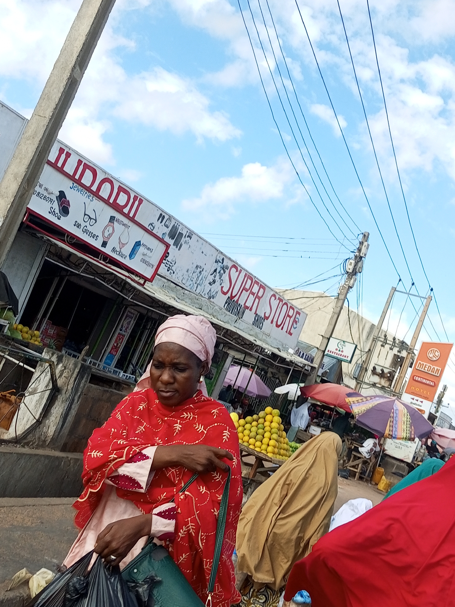 This is a medium-shot, eye-level, outdoor, street-level, slightly tilted shot of a bustling marketplace in Tempure, Nigeria, under a partly cloudy sky. The image is dominated by the presence of women dressed in traditional African attire. In the foreground, a woman wearing a red and white patterned kaftan and a pink headscarf, with gold hoop earrings, is holding a large black plastic bag filled with items, possibly purchases. Her gaze is directed downwards, and her hands are actively managing the bag and her clothing.
Behind her, other individuals are partially visible, including a person in a tan-colored garment and another in a bright red garment, suggesting more shoppers or vendors. Several market stalls are visible, featuring displays of fruit, with a prominent pyramid of yellow fruits, likely oranges or lemons, on a wooden table. Various umbrellas, some brightly colored, are set up to provide shade for the stalls, creating a vibrant and somewhat chaotic scene.
The background includes storefronts with signage. One prominent sign reads "SUPER STORE PROVISION SHOES." Another sign, angled and partially obscured, displays "ILODODI" with smaller text indicating "Jewelries, Shoe, Sun Glasses, Necklace, Perfumes, WRISTWATCH." Further down the street, other establishments are suggested by signage, including "MEDAN CONTINENTAL HOTEL." Power lines crisscross the sky, indicating the urban environment. The overall impression is that of a lively commercial hub in Tempure, Nigeria, with a strong focus on female shoppers and vendors.