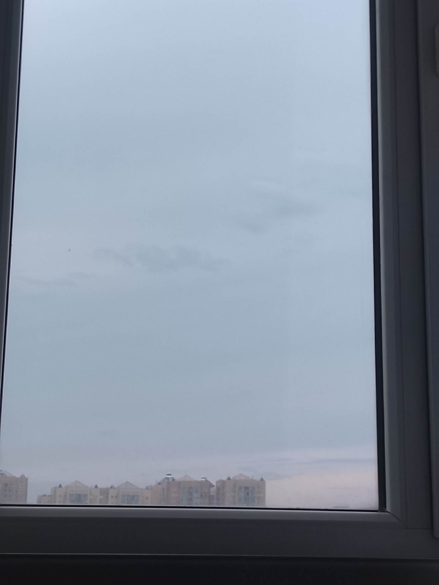 The image shows a view from a window, likely in Almaty, Kazakhstan, looking out at a hazy cityscape. The sky is overcast with a pale gray and white color. In the distance, a line of buildings is visible, appearing as a row of light-colored, multi-story structures with distinctive rooflines, possibly apartment complexes or administrative buildings. The lower part of the image is dominated by the dark frame of the window. A small dark speck, possibly a bird or an insect, is visible in the upper portion of the sky. The overall atmosphere is muted due to the cloudy weather.