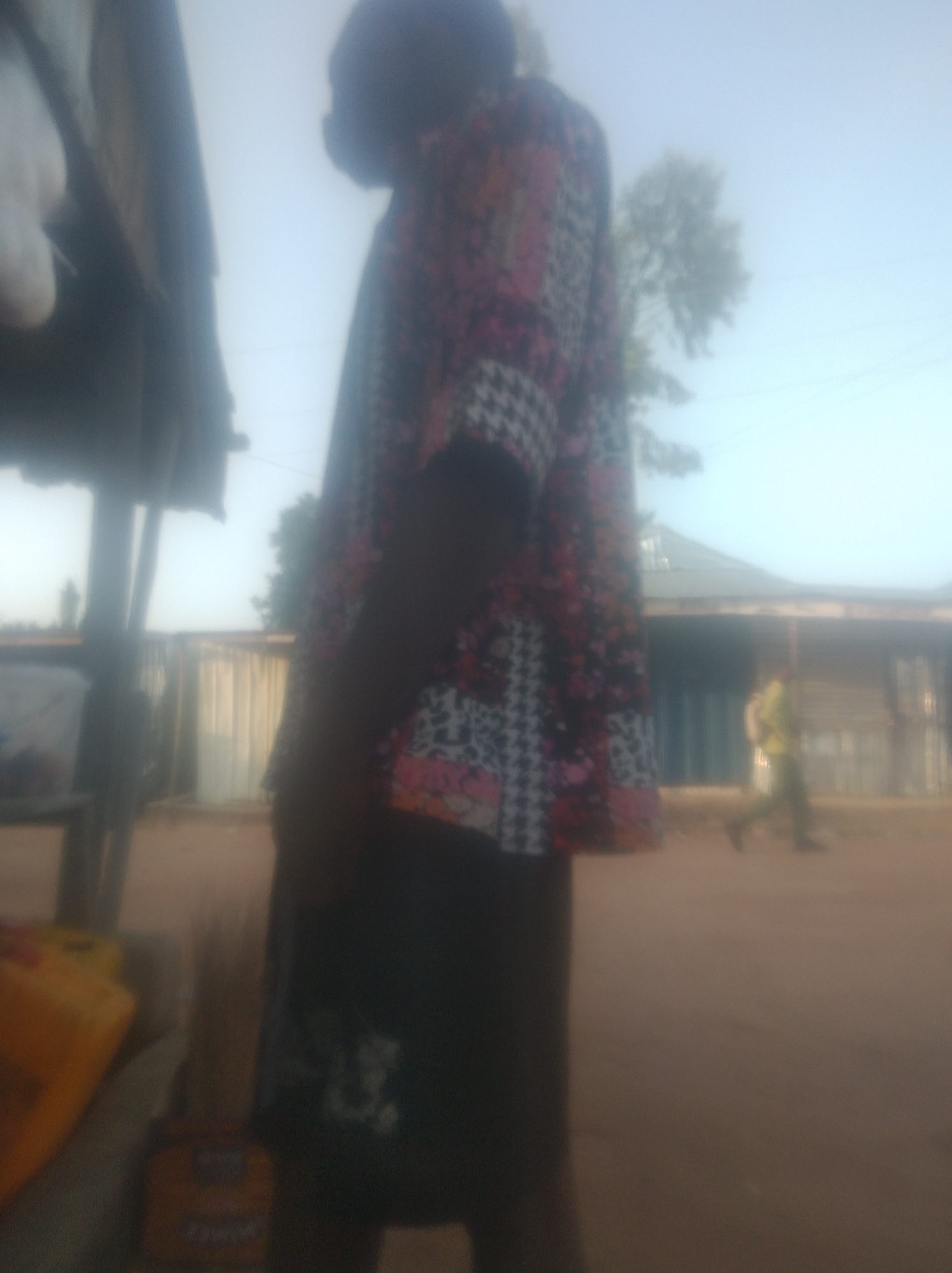 This is a slightly out-of-focus, medium-shot photograph taken outdoors, likely in a dusty, unpaved area of Jalingo, Nigeria. The scene is dominated by a person in the foreground, viewed from the side, wearing a brightly colored, patterned shirt and dark pants. The shirt features a complex geometric design with pink, black, white, and orange elements. To the left of the foreground person, there is a partial view of a structure with a dark, sloped roof supported by posts, possibly a stall or shelter. In the background, another person is walking away from the camera, wearing a light-colored shirt and dark pants, and carrying a backpack. Behind the walking person, there is a long, low building with a light-colored facade, possibly a market or shop. The ground appears to be dirt or sand. The sky is overcast and hazy, suggesting it could be early morning or late afternoon, or a dusty atmospheric condition.