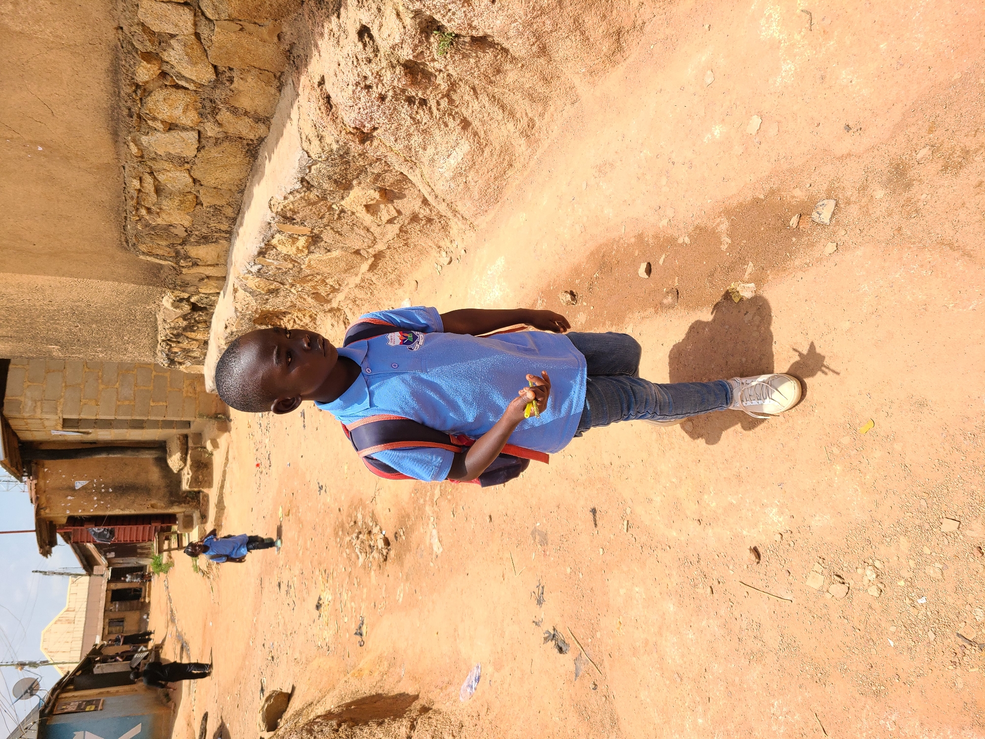A young boy stands on a dirt road in an urban setting. He wears a light blue short-sleeved shirt, jeans, and white sneakers. A backpack with red straps is slung over his shoulder. He is holding a small yellow object in his right hand. Behind him are buildings constructed of brick and other materials. The dirt road shows signs of wear and some scattered debris. Another person, wearing a blue shirt, is slightly out of focus in the background, further down the road. The location appears to be a residential area.