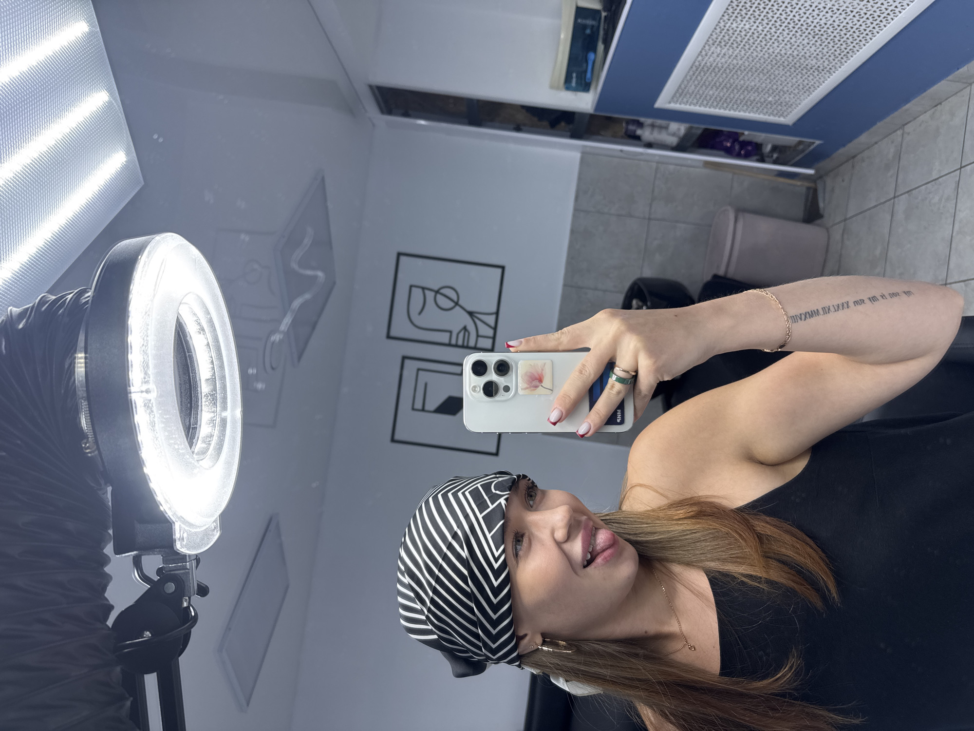 A young woman with blonde hair, wearing a black tank top and a black and white patterned bandana, takes a selfie in a mirror. She is sticking her tongue out and looking to the left. She has a gold necklace, hoop earrings, a green ring, and a thin gold bracelet. Her fingernails are painted red with white tips. In the background, there are framed abstract art pieces, tiled walls, a trash can, and shelves with various bottles and containers. A ring light is visible above her, and a large fluorescent light fixture is on the ceiling. The scene appears to be in an indoor salon or studio, possibly in Chaikovsky, Russia.
