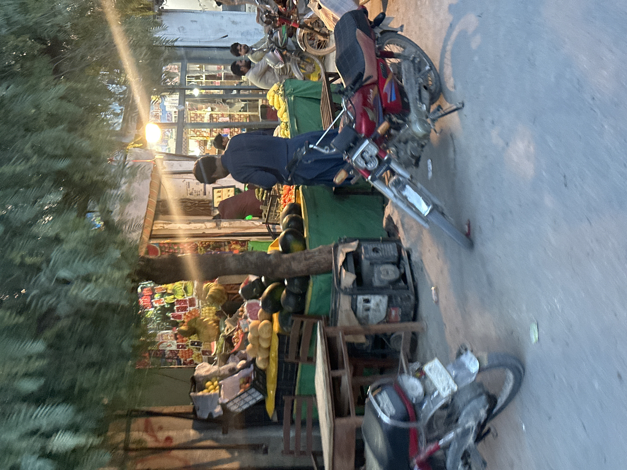 This image captures an evening or nighttime scene at a street market or roadside fruit stall. The overall setting is an outdoor area alongside a road, illuminated by artificial light sources.
Several subjects are visible. In the foreground, a red motorcycle with a black seat is prominently parked on the asphalt road, facing towards the right side of the frame. Another motorcycle is partially visible in the bottom right corner. A large, leafy tree occupies the left side of the image, with its dense foliage extending high up.
Towards the right and center, a fruit and vegetable stall is set up, displaying a variety of produce. Bright yellow fruits, possibly bananas or corn, are stacked on a green tarp covering a display stand. Watermelons and other colorful fruits and vegetables, including what appear to be oranges and possibly tomatoes, are arranged in crates and on shelves. Behind the stall, a shopfront with illuminated interior goods is visible through its windows.
Several people are present, indicating ongoing activity. A man wearing a dark blue tunic (shalwar kameez style) is standing near the red motorcycle, seemingly looking towards the fruit stall. Further back, another man in a light-colored tunic is visible near the stall. Other figures are vaguely discernible behind the main stall area.
A portable generator is placed on a wooden stand near the fruit display, suggesting its use for powering lights or other equipment at the stall. The scene implies shopping or trading activities taking place under artificial light. The time of day is clearly evening or night due to the reliance on electric lighting, which casts shadows across the street and objects. No legible text is visible in the image.