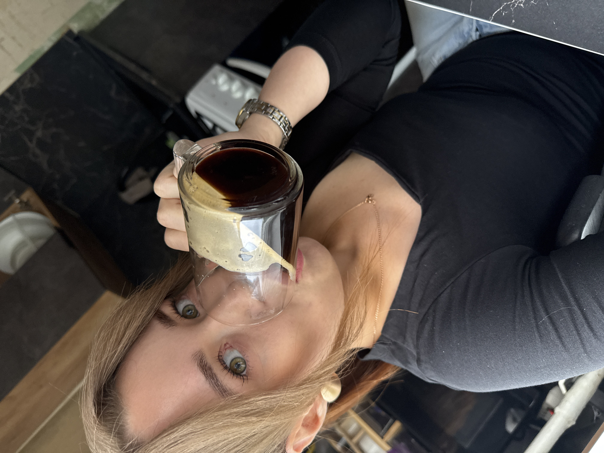 A young woman with blonde hair is drinking coffee from a clear mug. She is wearing a black, long-sleeved shirt and light-colored pants or shorts. She has light green eyes and is wearing simple stud earrings. The background appears to be a kitchen or dining area, with dark countertops and a white appliance visible. There are no specific landmarks in the image to indicate the city of Chaikovsky, Russia.