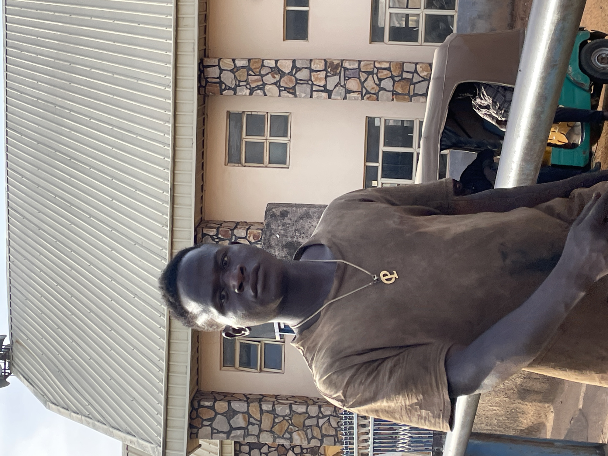 The image captures a young man in what appears to be an outdoor, urban setting in Jos, Nigeria. The man, with dark skin and short dark hair, is positioned in the mid-ground, slightly to the right of the frame, looking directly towards the viewer with a serious or neutral expression. He wears a plain, light brown or tan short-sleeved t-shirt, which appears somewhat dusty or worn. Around his neck, he wears a gold-colored chain with a distinctive pendant shaped like a stylized letter "e" or similar symbol. His right arm is visible, bent at the elbow, and he appears to be resting or leaning on a prominent silvery metal railing that runs horizontally across the lower part of the image.
Behind the man and to his left stands a multi-story building with light-colored, possibly cream or beige, walls. The building features decorative bands of natural stone masonry with irregular gray, brown, and reddish stones, particularly noticeable between floors. Numerous windows with white frames and dark panes are visible on its facade. The upper part of the building, on the left side of the frame, is topped with a light gray corrugated metal roof, featuring parallel diagonal ridges.
To the far right, partially obscured by the railing and the man, a section of a light blue-green, older model vehicle, possibly a utility truck, is visible, including a wheel. The lighting suggests bright daylight, likely midday or early afternoon, casting clear shadows. The overall scene conveys a candid moment in a bustling environment. No other visible text is present apart from the necklace pendant.